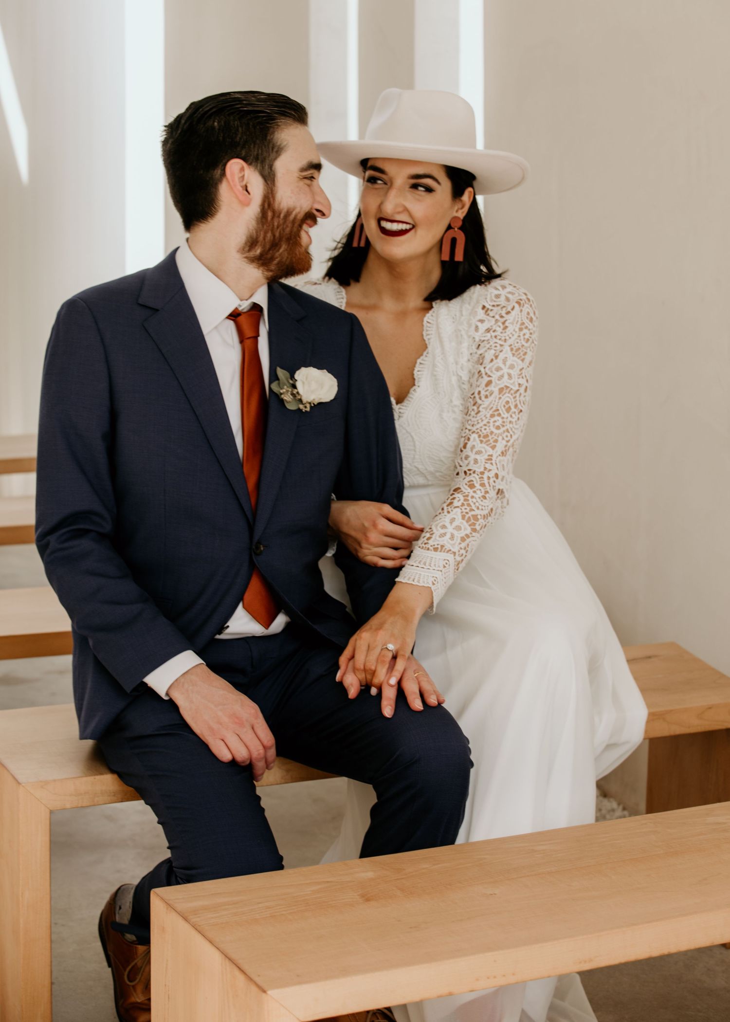 Bride in lace dress and white cowboy hat sitting with groom on a wooden bench, laughing together.