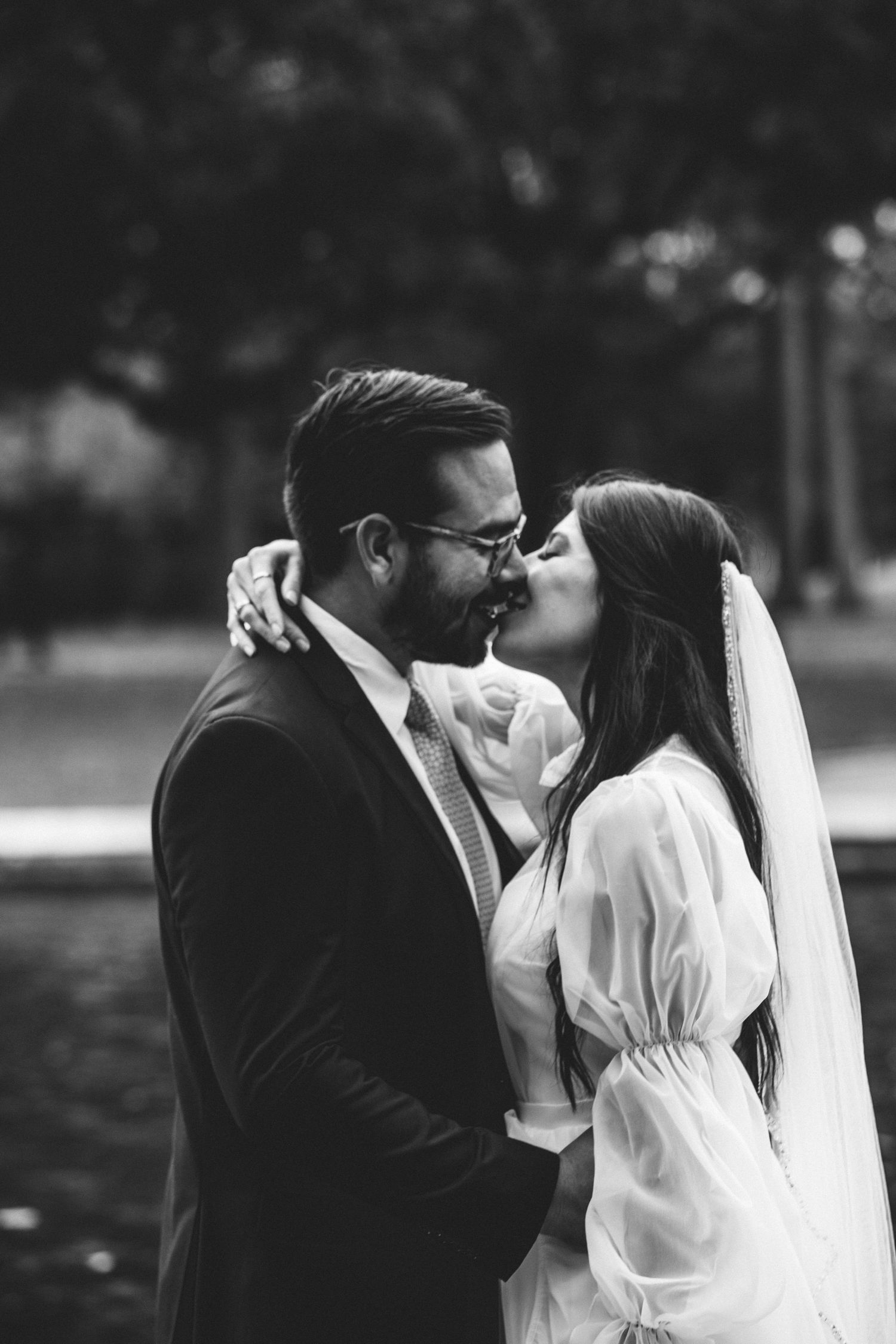 Black and white portrait of bride and groom sharing a romantic kiss outdoors near water.