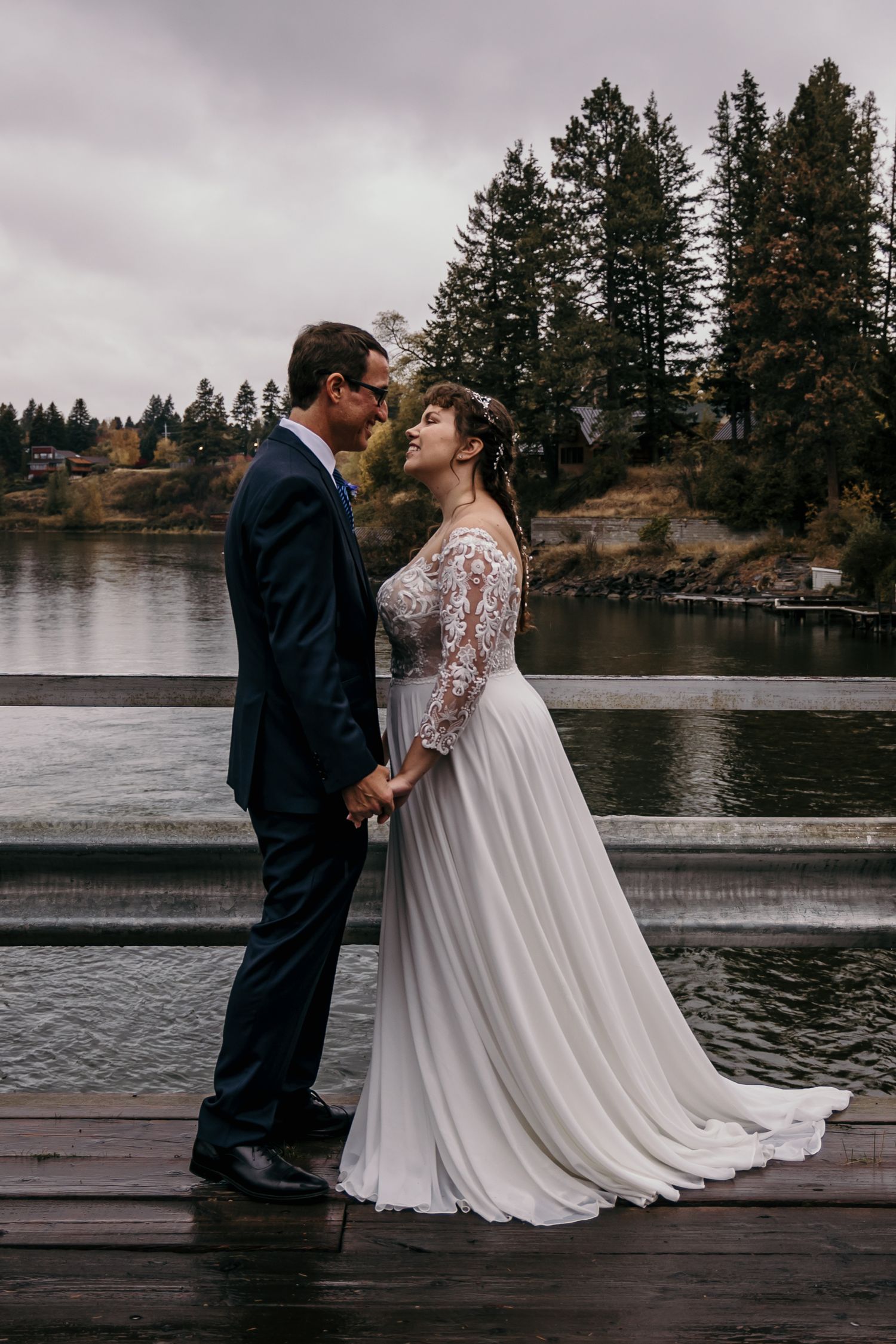 Bride in elegant lace gown and groom in navy suit hold hands facing each other on a lakeside dock.