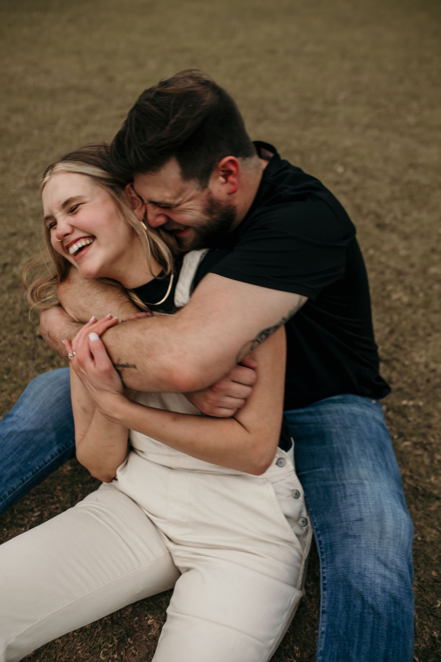 Man embracing laughing woman from behind while sitting on grass outdoors.