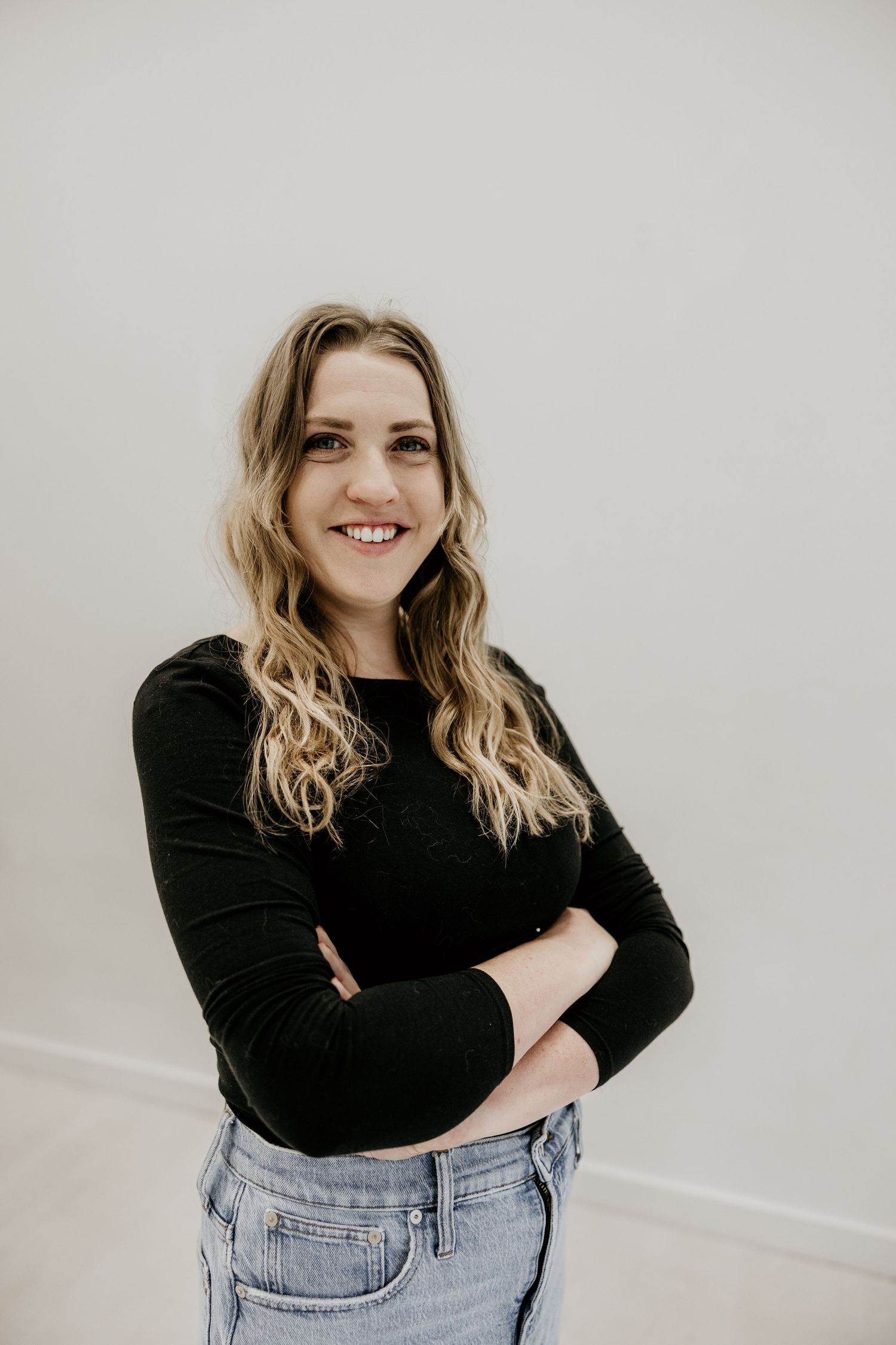 Smiling woman in black long sleeve top and jeans with arms crossed against a light wall.