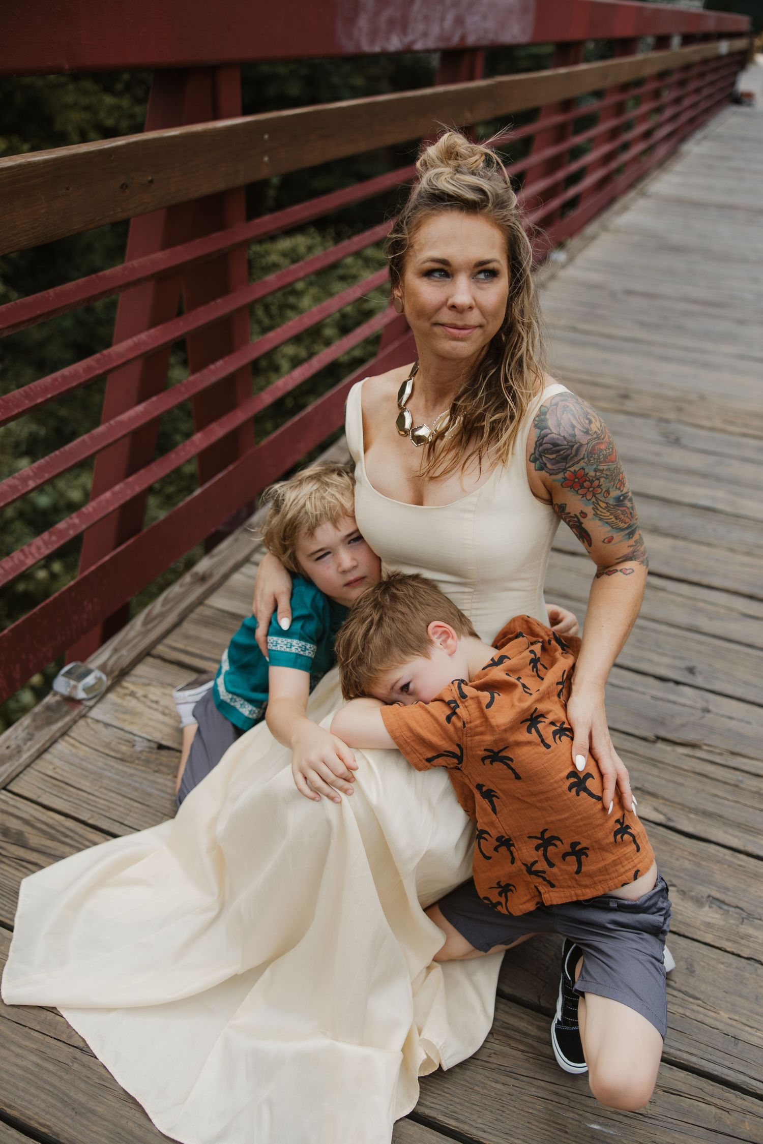 A tattooed mother in a cream dress sits on a wooden bridge hugging her two young sons.