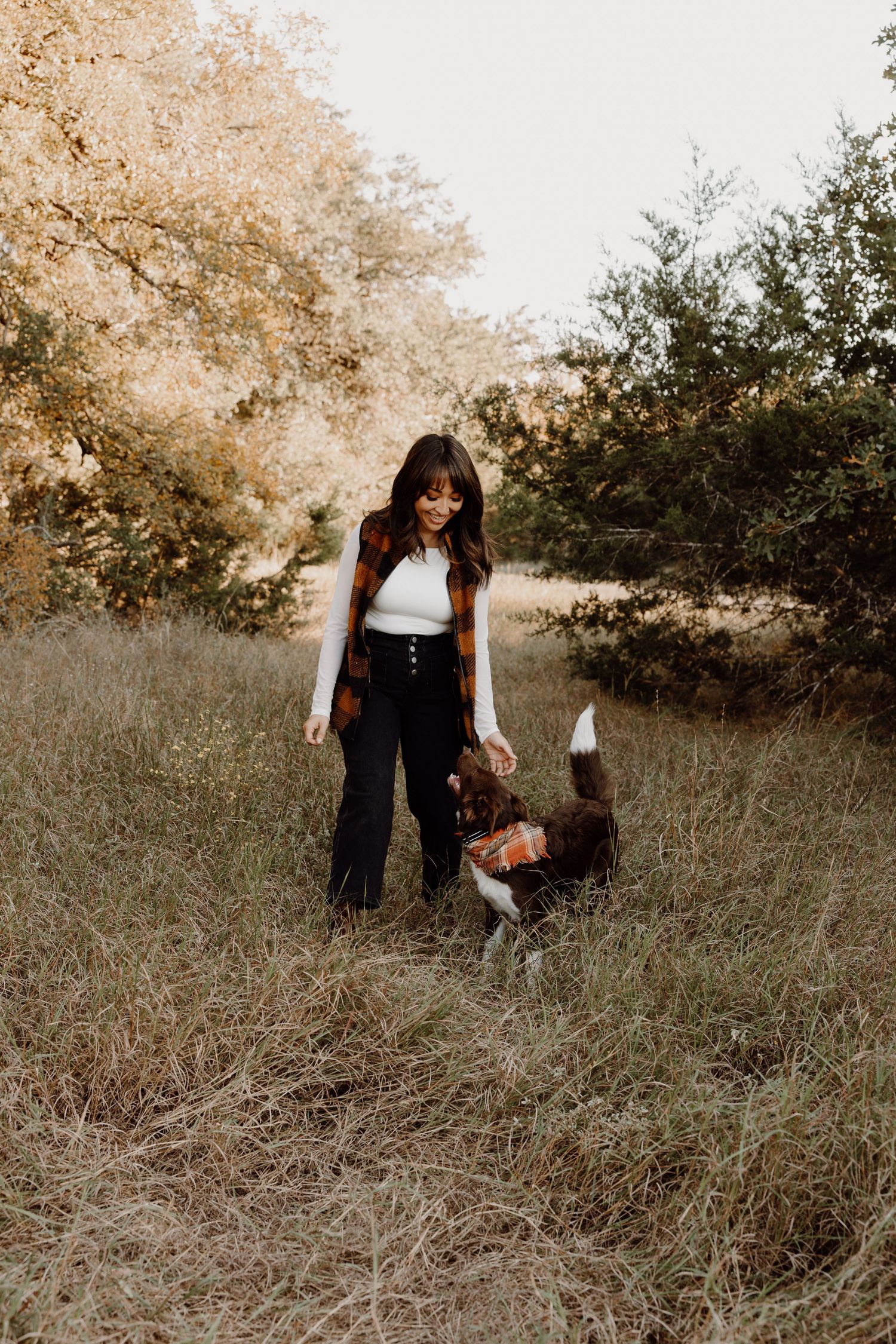 Young woman in plaid vest playing with a brown and white dog in an open grassy field.