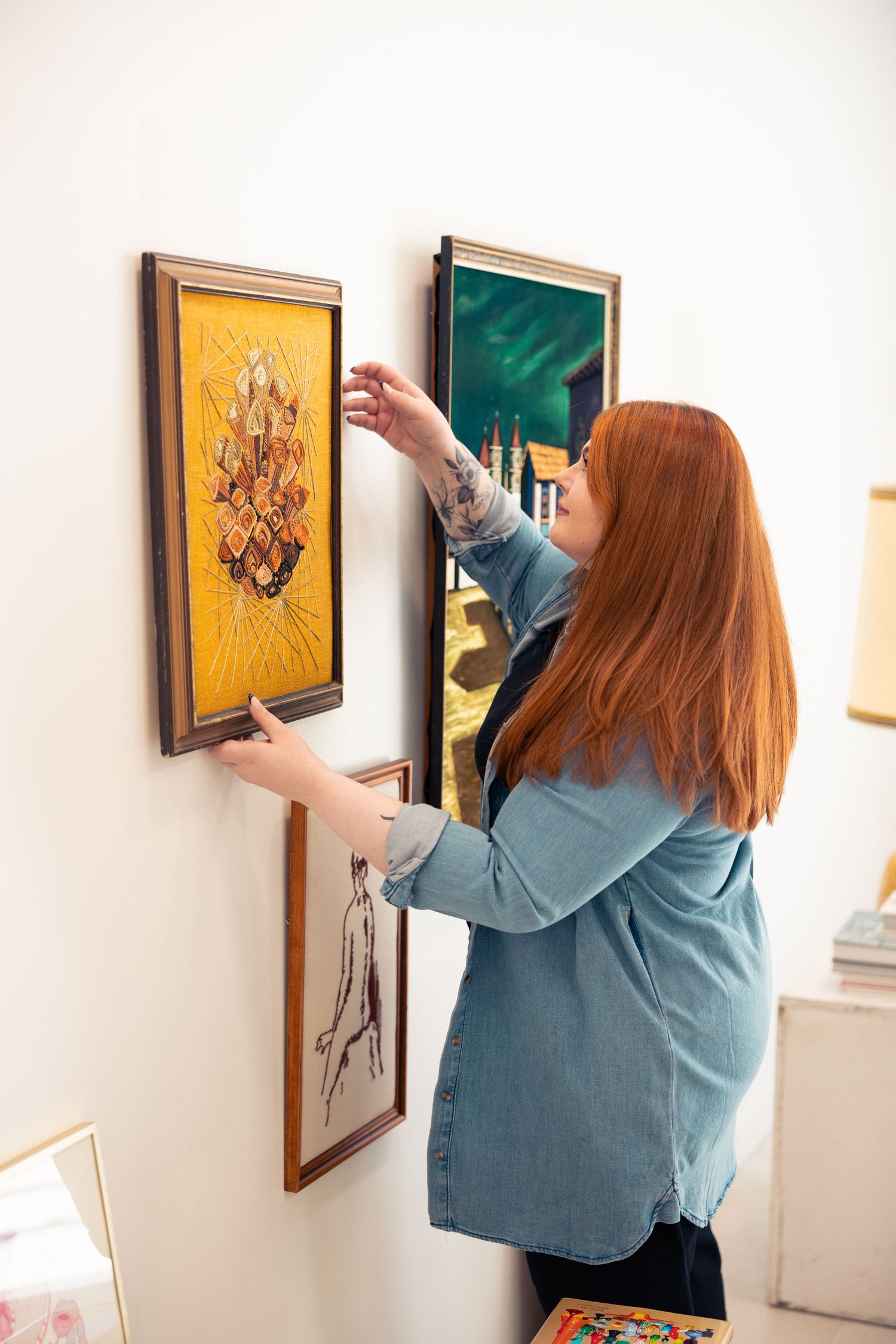 Redheaded woman arranging framed vintage artwork on white wall in bright interior space.