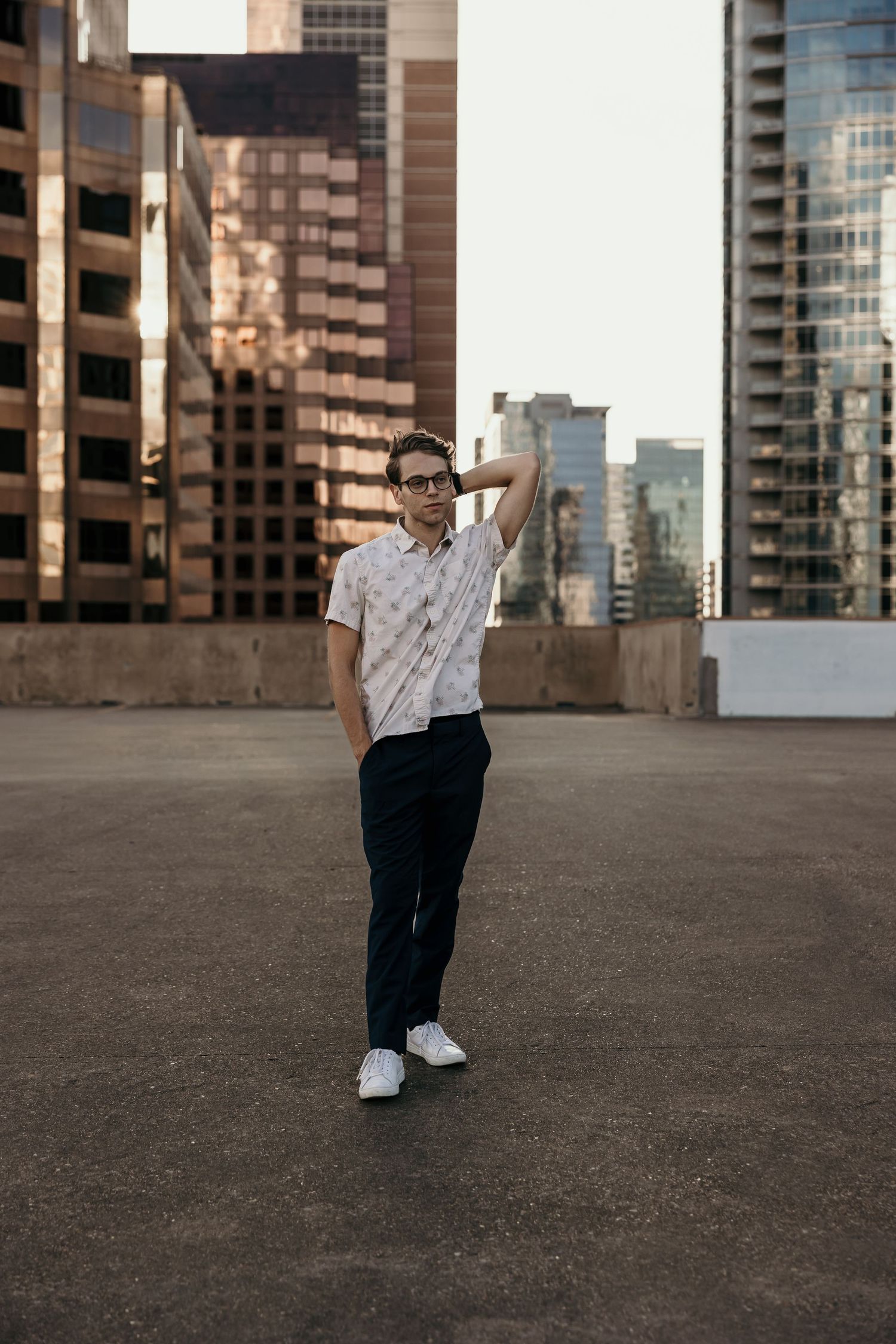 Young man in patterned shirt and dark pants poses confidently on urban rooftop surrounded by city skyscrapers.