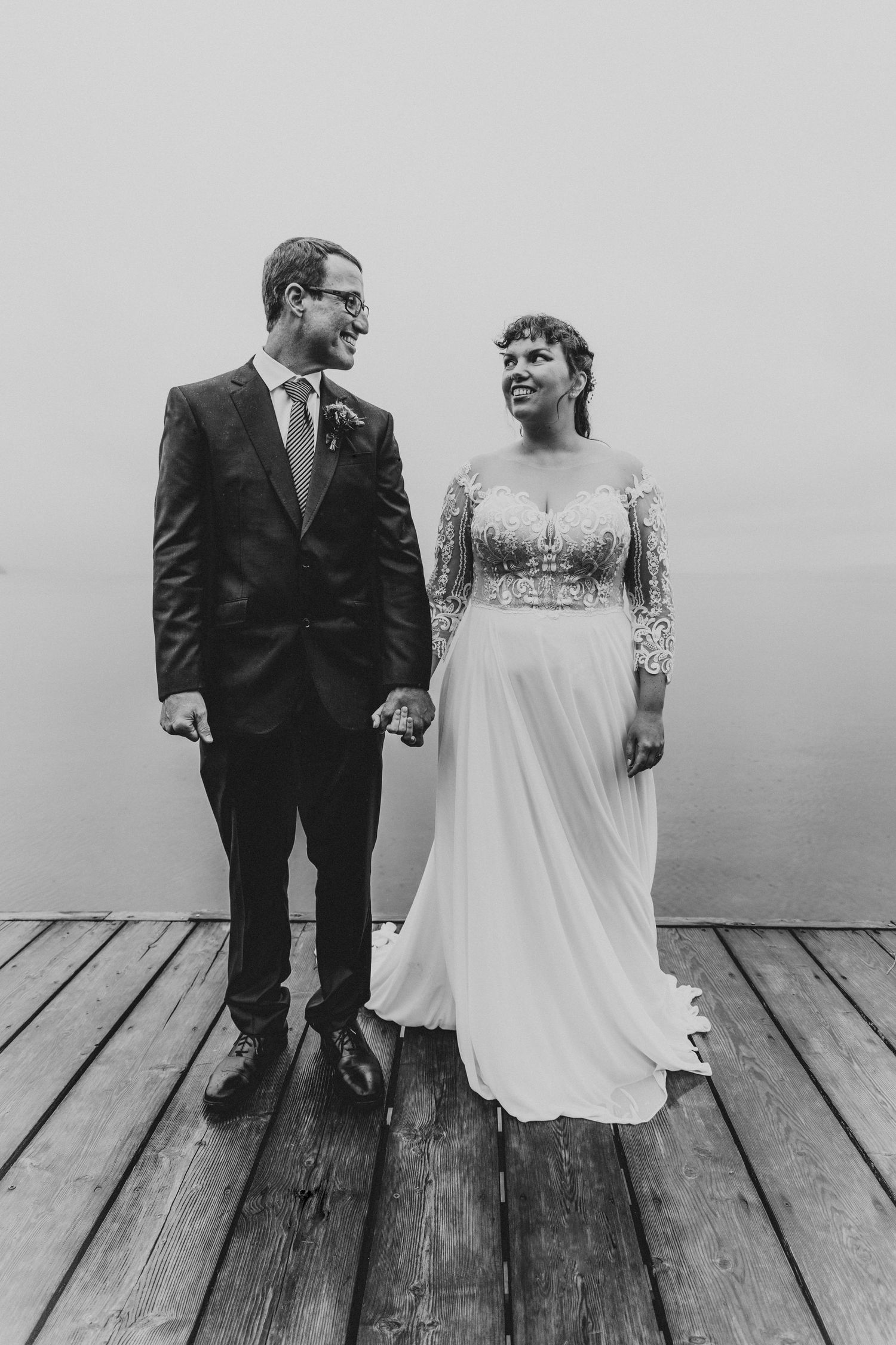 Black and white photo of smiling bride and groom holding hands on a wooden dock by the water.