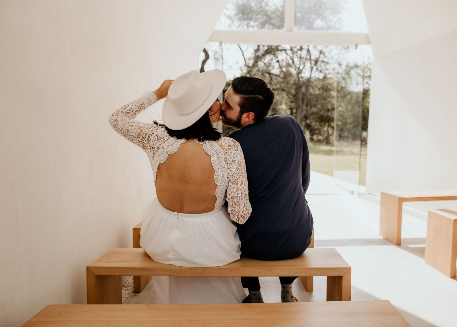 Couple in wedding attire sitting on a wooden bench, sharing a kiss, bride in lace dress holding white hat.