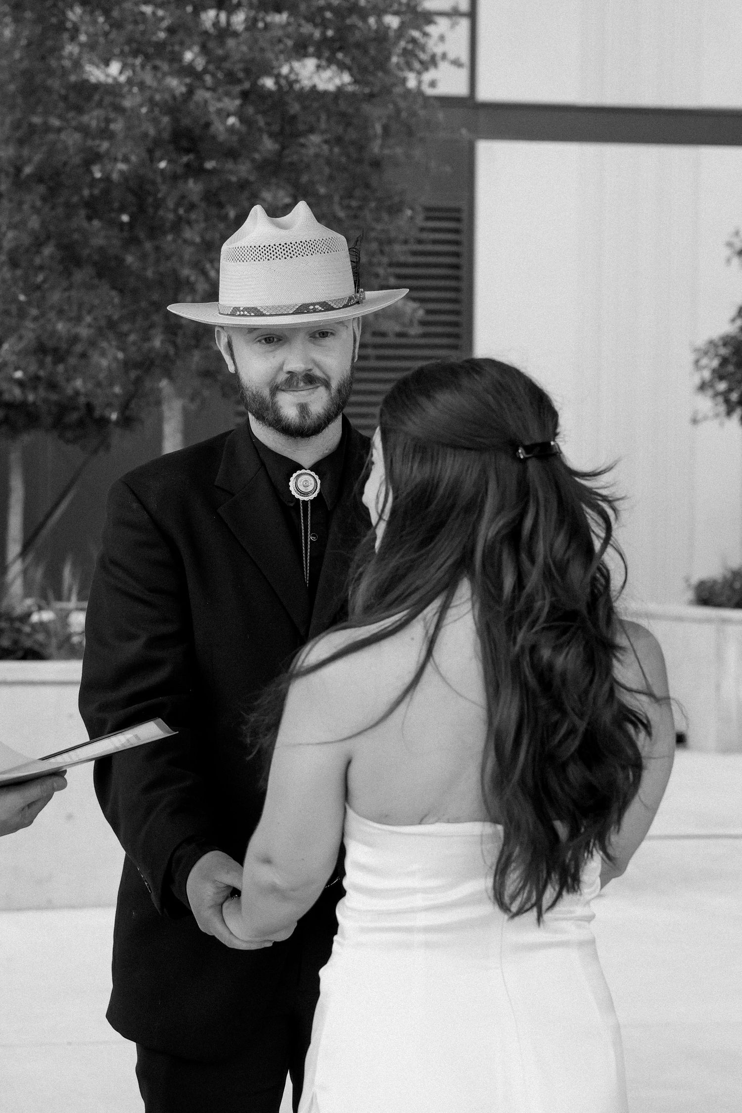 Black and white image of bearded groom in cowboy hat reading vows to bride at wedding ceremony.