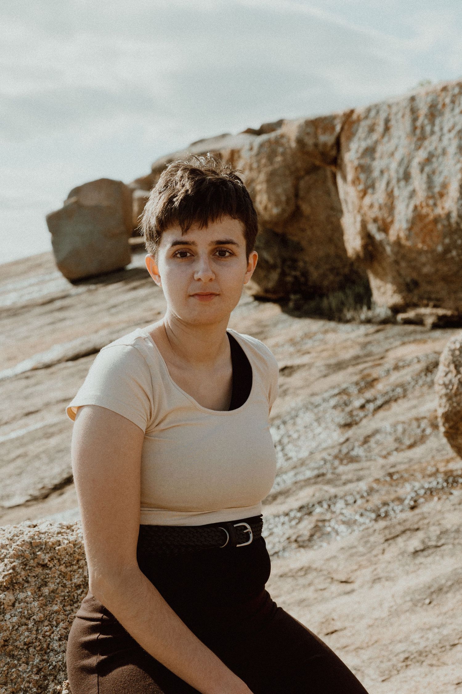 Person in beige top and dark belted pants sitting on rocks with boulders behind.