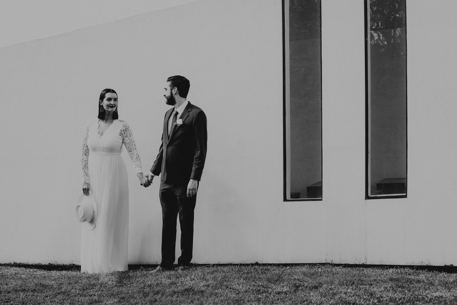Black and white photo of bride and groom holding hands beside a modern white chapel wall.