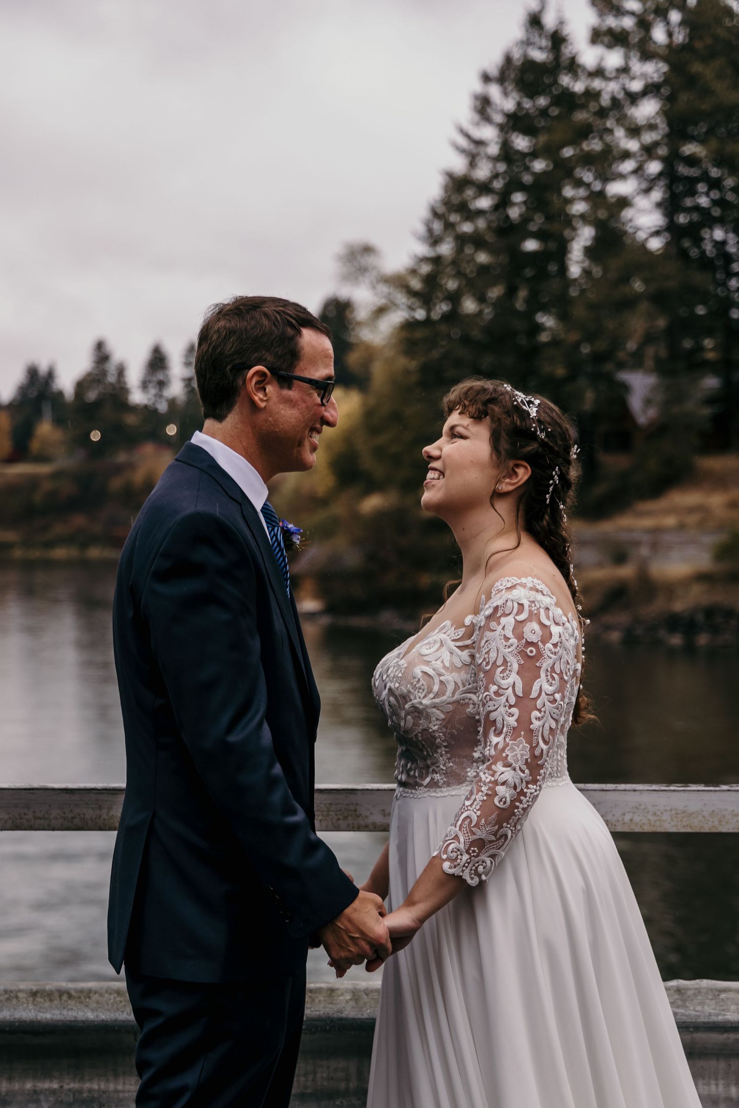 Bride in lace wedding gown laughs joyfully while holding hands with groom on a misty lakeside bridge.