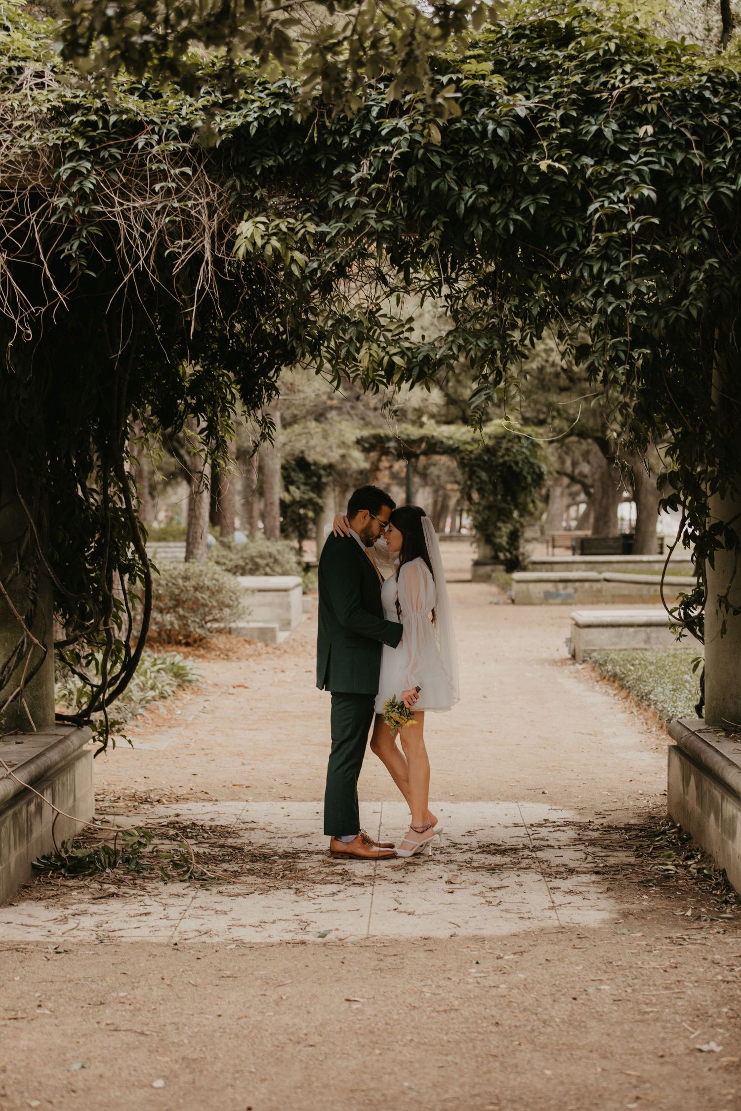 Bride and groom embrace on a garden pathway framed by lush green ivy-covered archway.