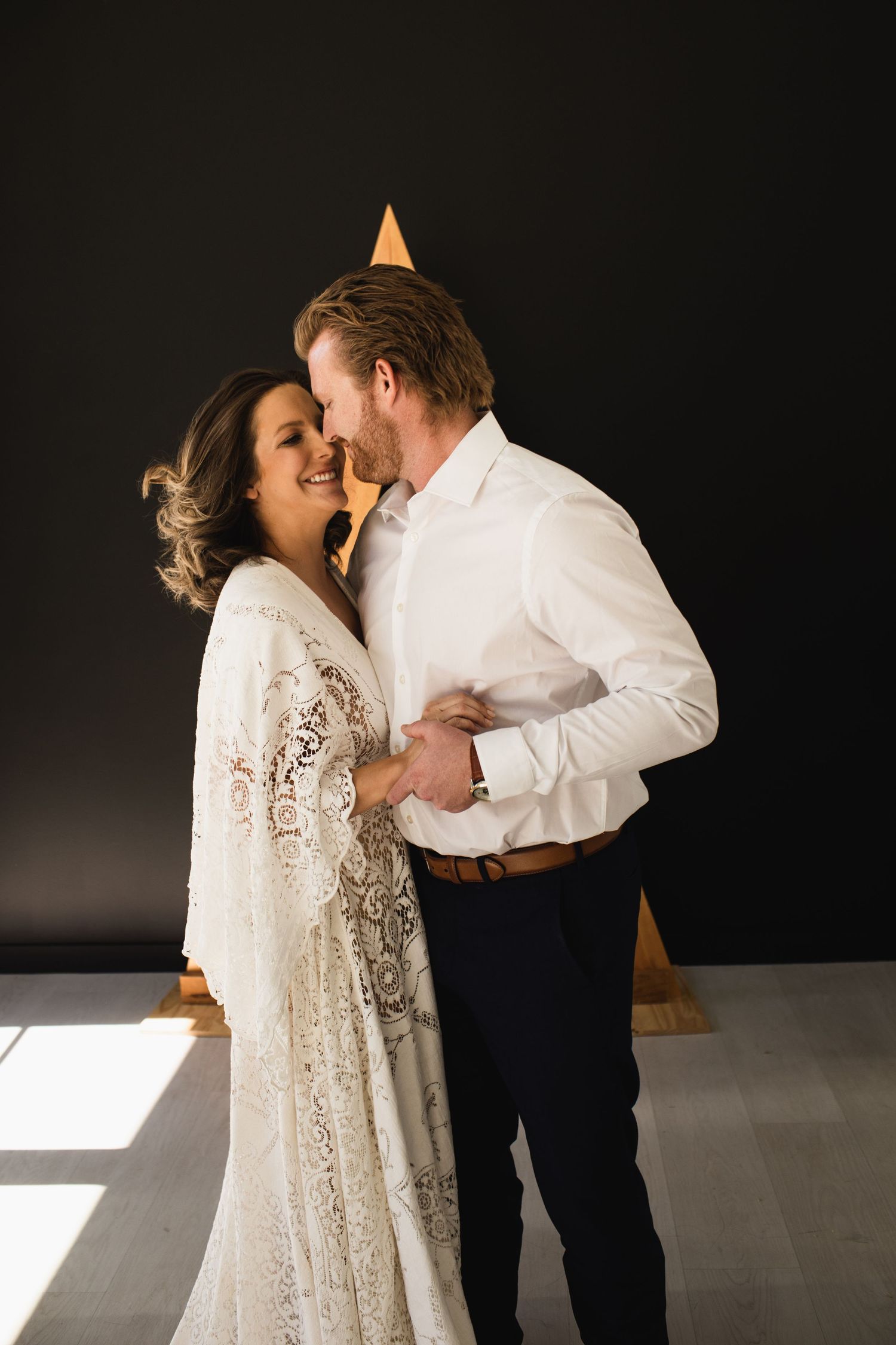 Couple in wedding attire sharing a romantic moment, bride in lace gown, groom in white shirt, dark background.