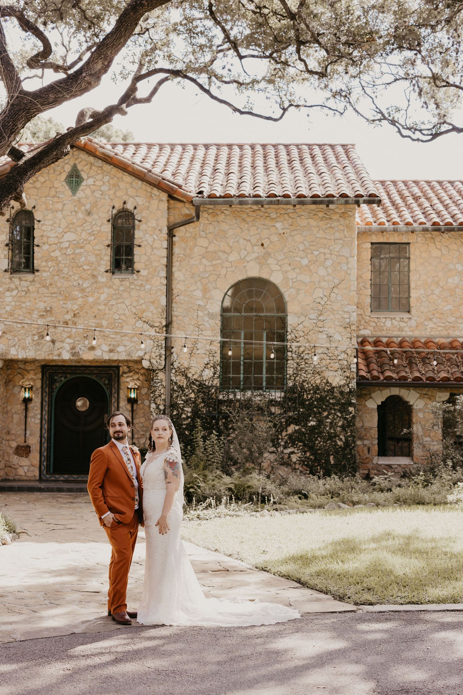 Bride and groom pose before a rustic Spanish-style stone villa with red tile roof.