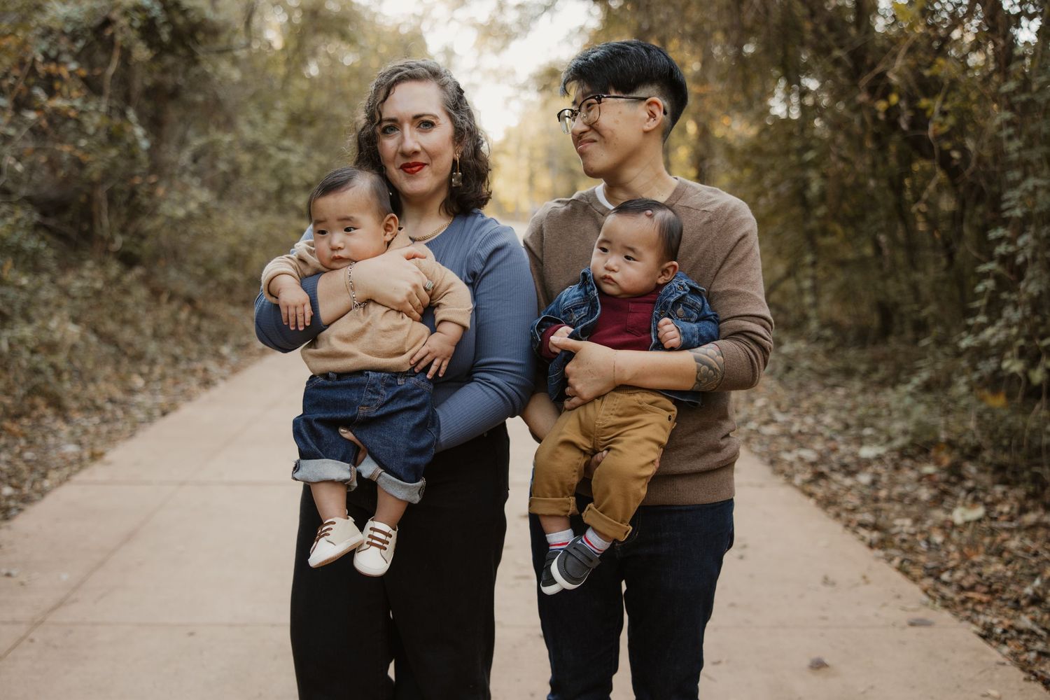 Two parents each holding a baby boy standing on a tree-lined path in autumn foliage.