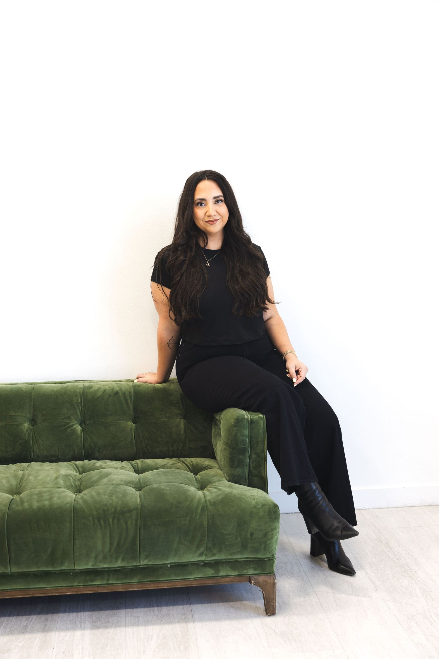 Woman in black outfit sitting on green velvet tufted sofa against white wall.