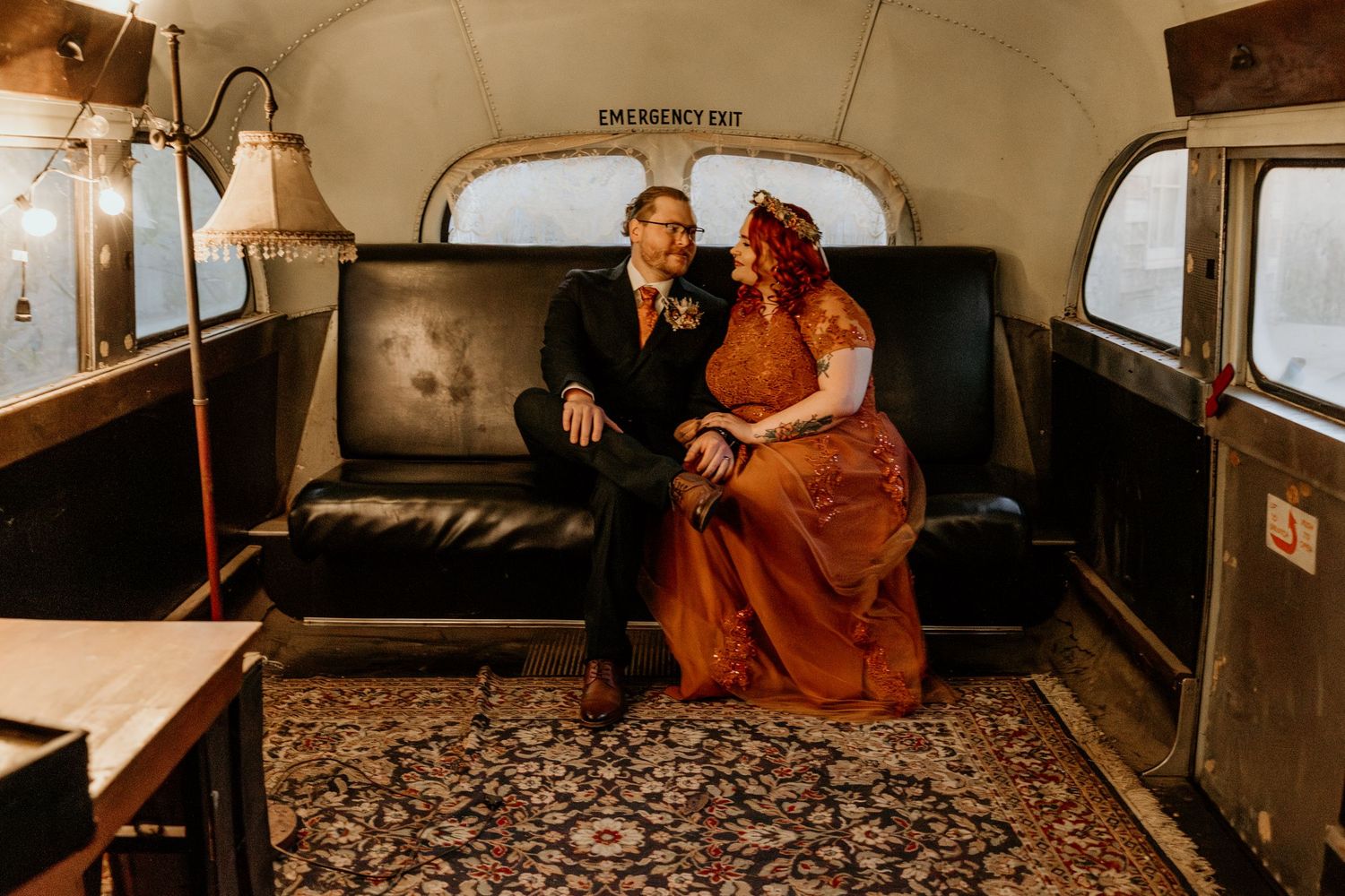 Bride in rust orange gown and groom sit together on vintage sofa inside a classic Airstream trailer.