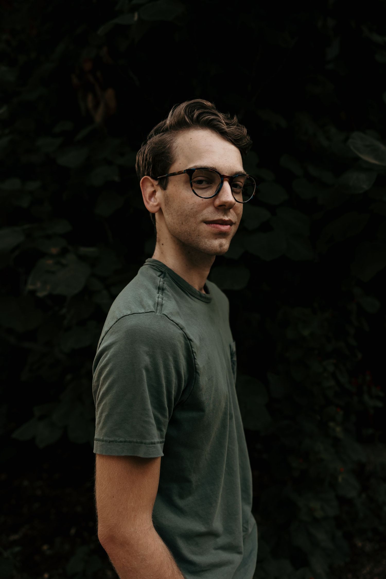 Young man in glasses and olive green t-shirt poses against a dark leafy background outdoors.