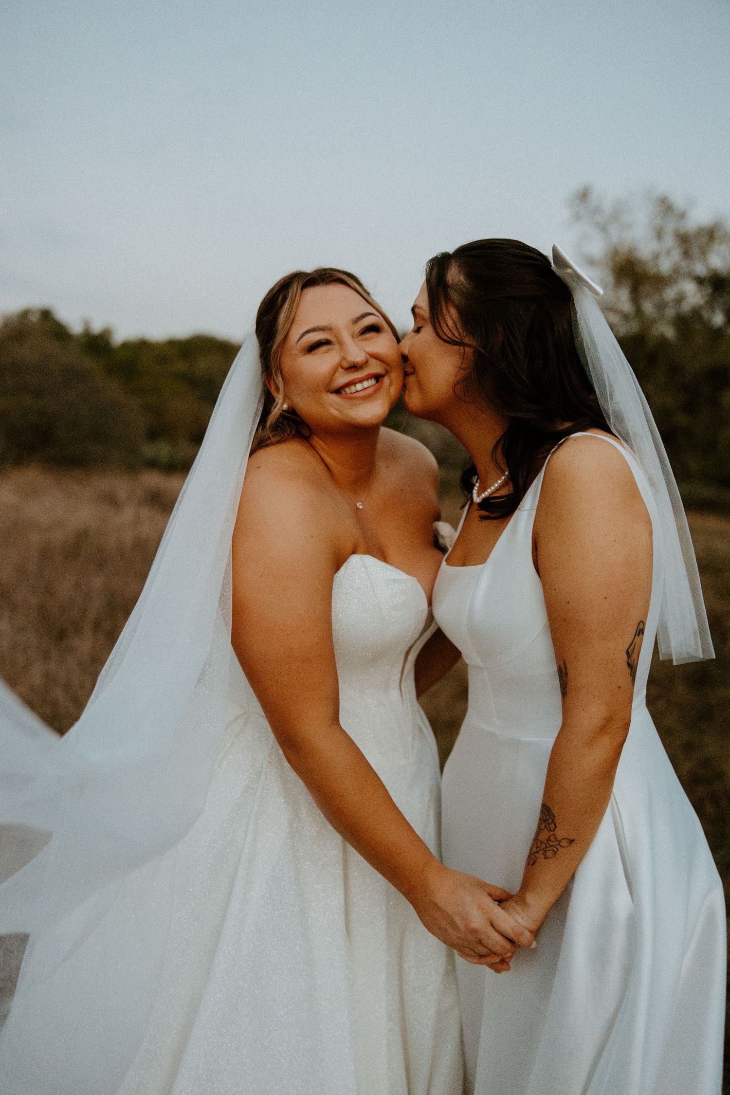 Two brides in white wedding gowns holding hands, one kissing the other on the cheek outdoors.