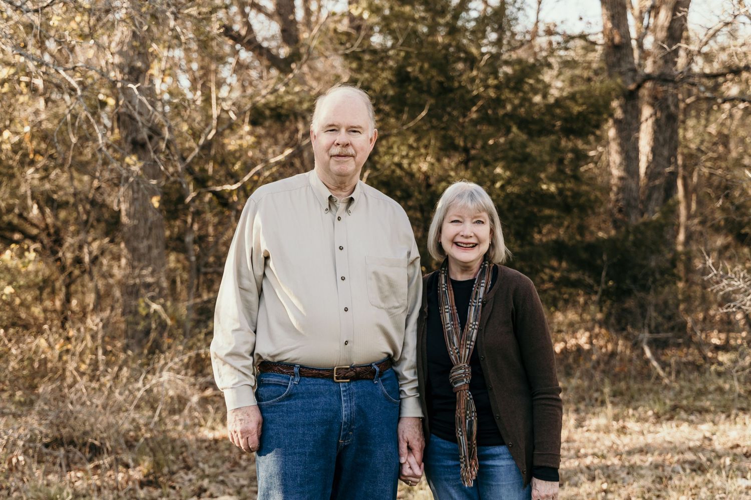 Smiling older couple standing together in a wooded outdoor setting during autumn season.
