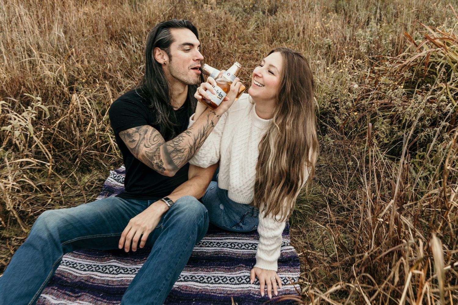 Tattooed man playfully biting a flower while woman laughs beside him on a blanket in a field.
