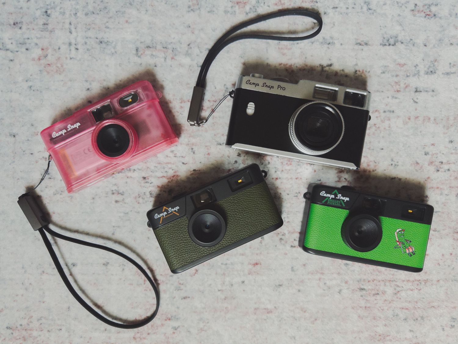 Four colorful vintage-style film cameras in pink, black, olive green, and green with wrist straps on a white surface.