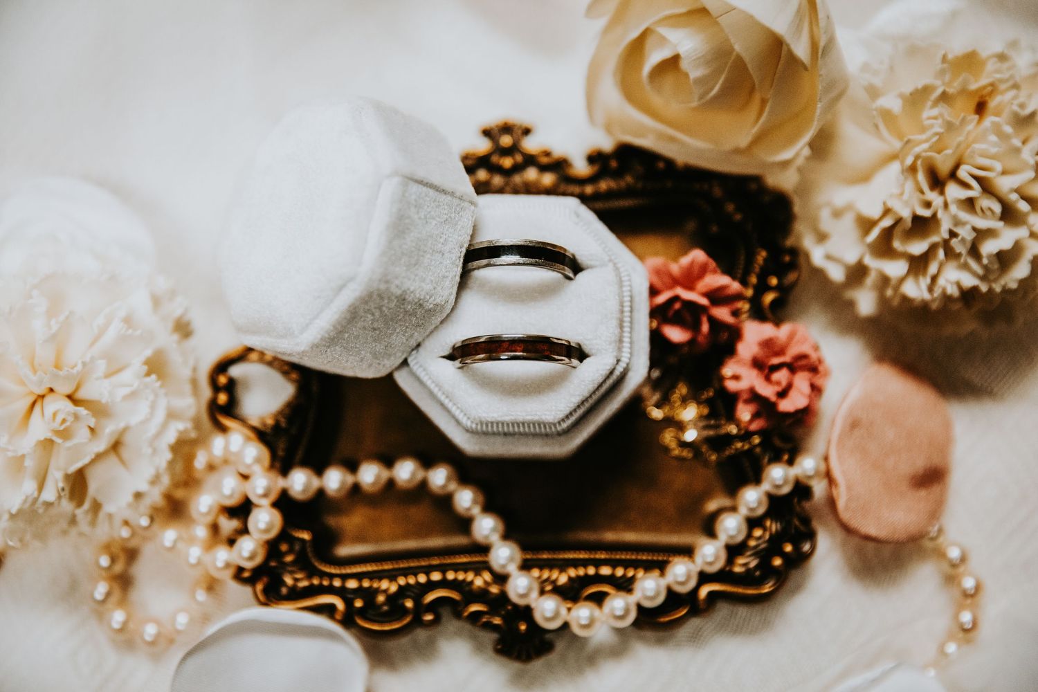 Two gold wedding bands displayed in a velvet ring box surrounded by pearl necklace and delicate flowers.