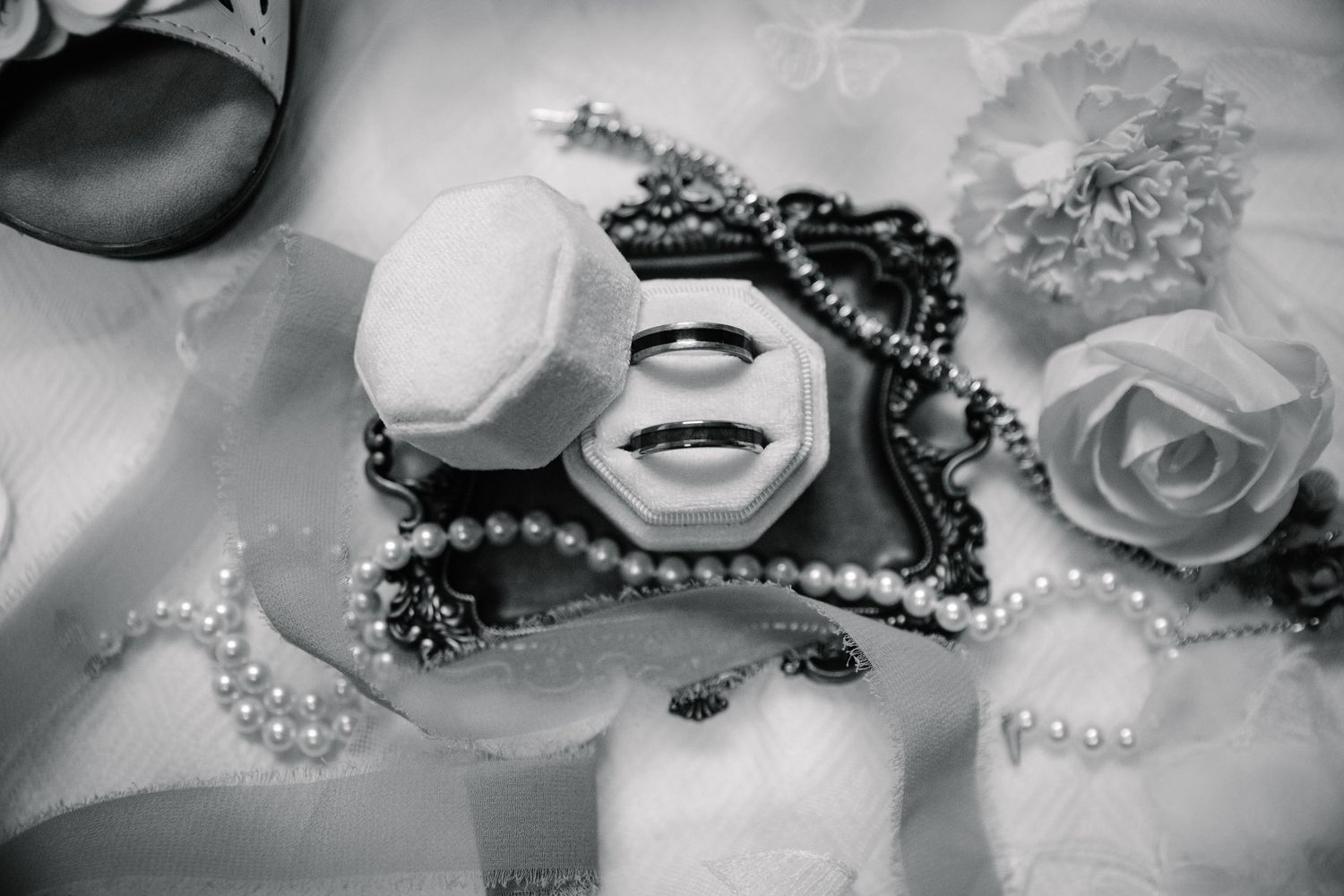 Black and white photo of wedding rings in a velvet box surrounded by pearl necklaces and floral decorations.