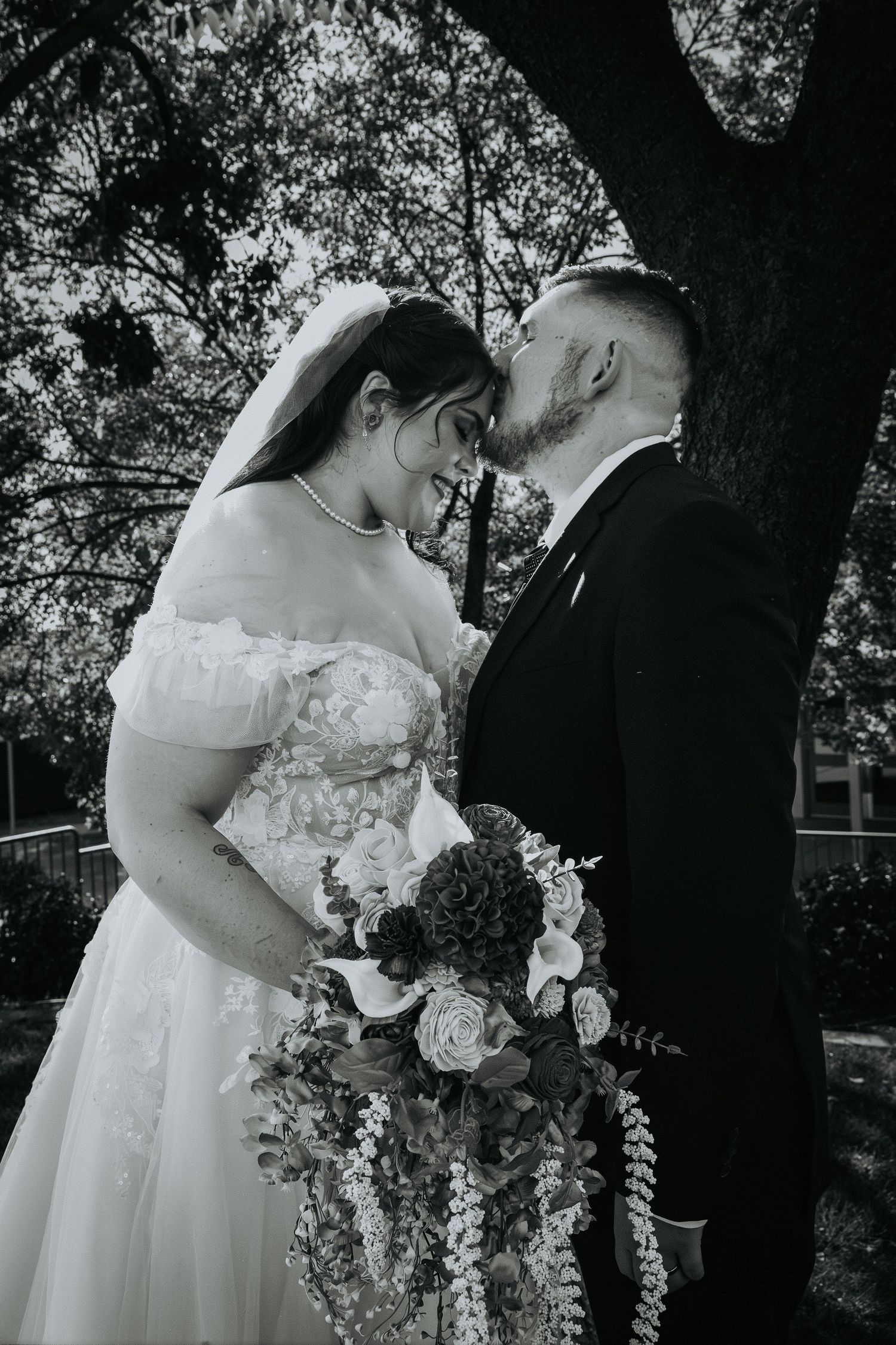 Black and white photo of a bride and groom sharing a romantic moment, bride holding a floral bouquet outdoors.