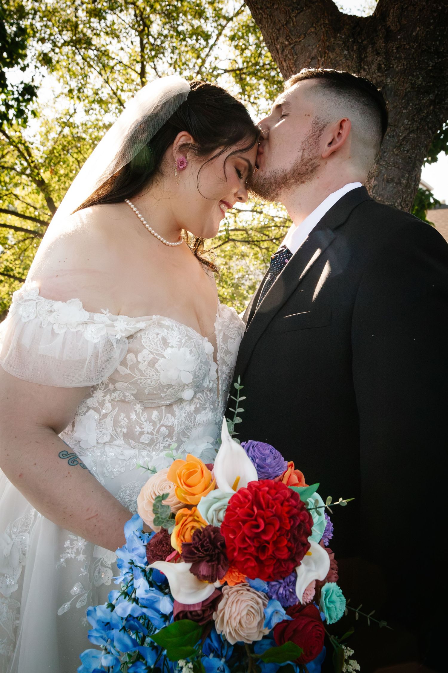 Bride and groom sharing a tender kiss outdoors, bride holding a vibrant bouquet of red, orange, blue, and white flowers.
