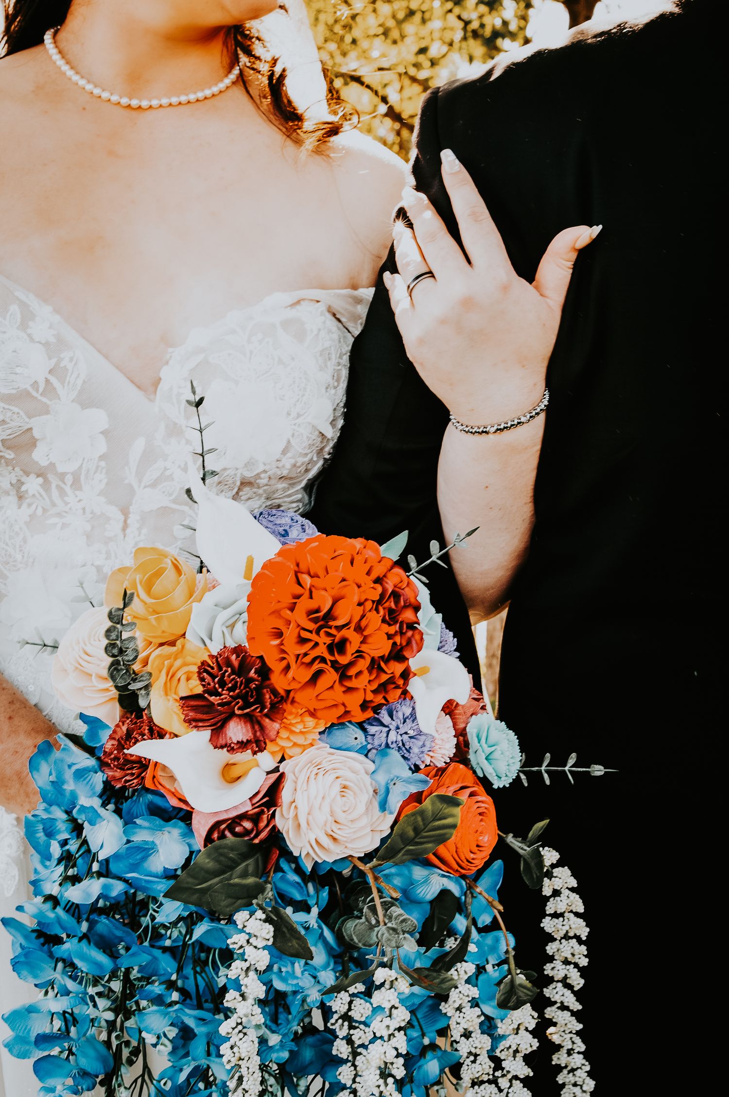 Bride in lace gown holding vibrant bouquet with orange dahlias, blue flowers, and roses beside partner in black.