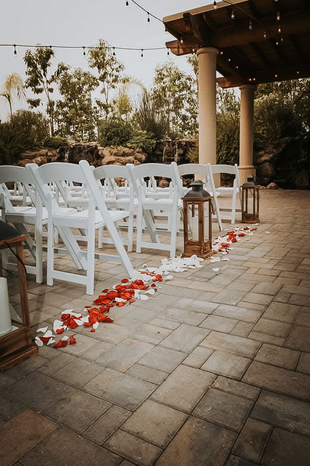 An outdoor wedding ceremony setup with white chairs, lanterns, and rose petals on the ground.