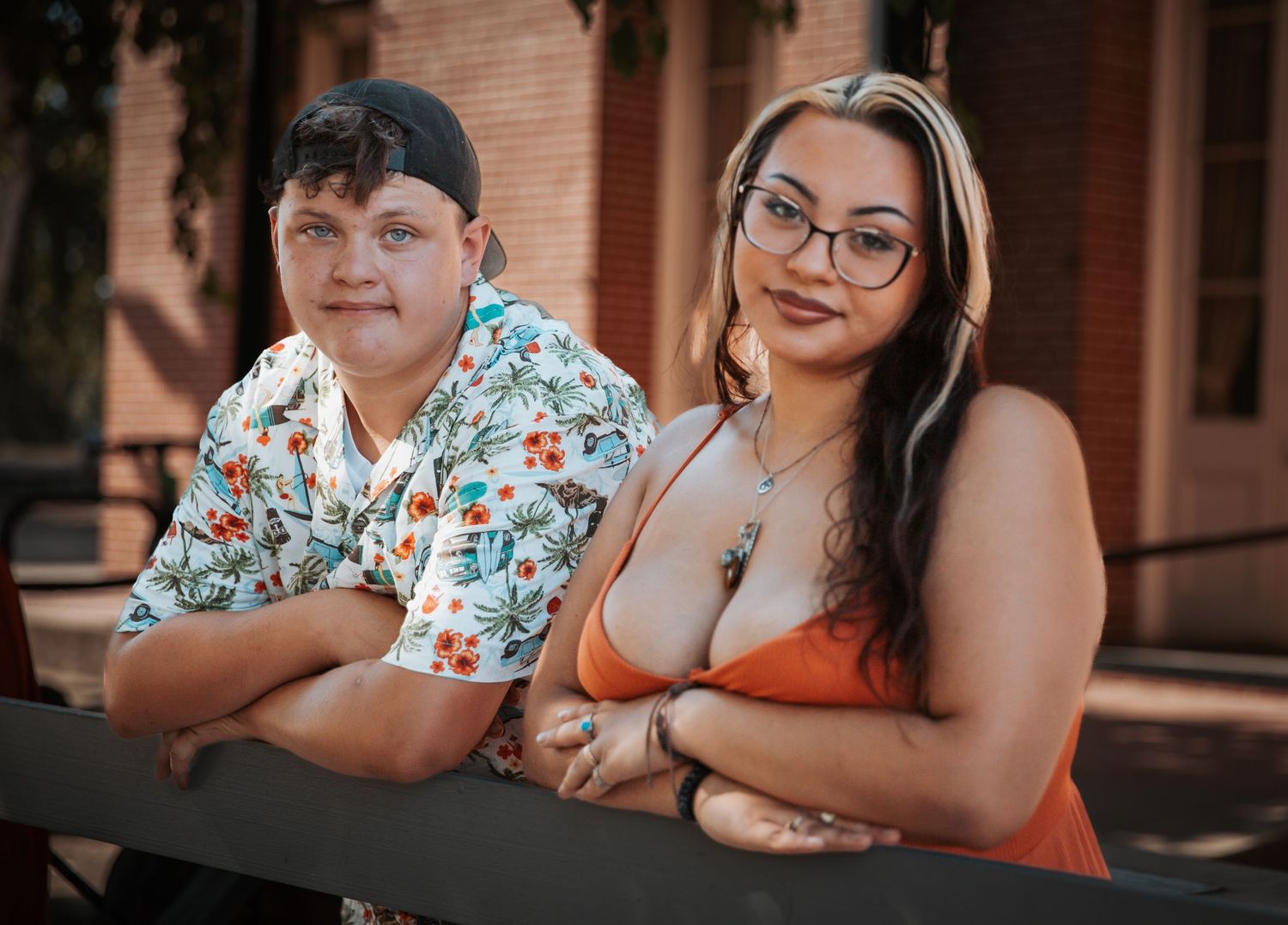 Two young people pose together outdoors near a brick building, wearing casual summer attire.