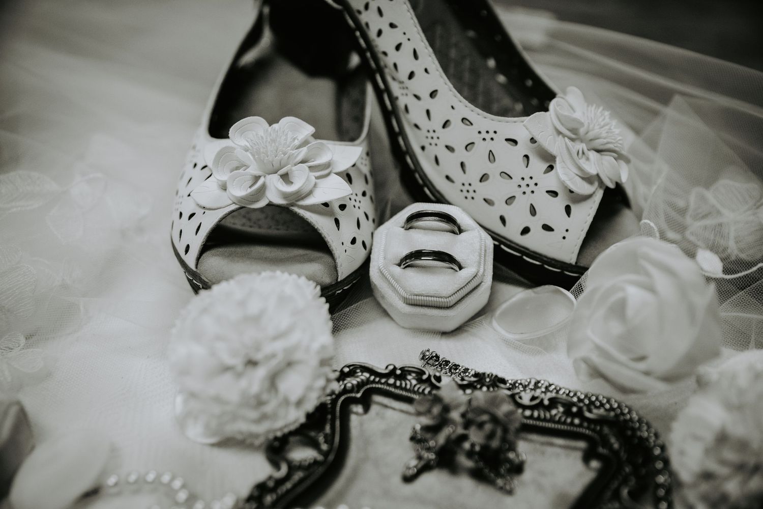 Black and white photo of elegant bridal shoes with floral details, wedding rings, and decorative accessories arranged beautifully.