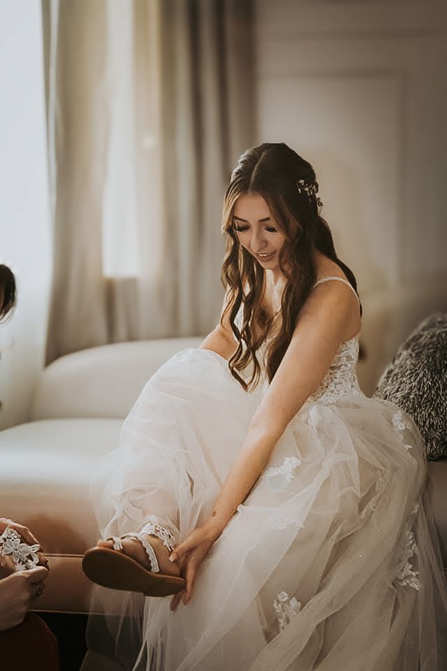 A woman in a white wedding dress sits on a couch, applying her makeup and getting ready for her wedding day.