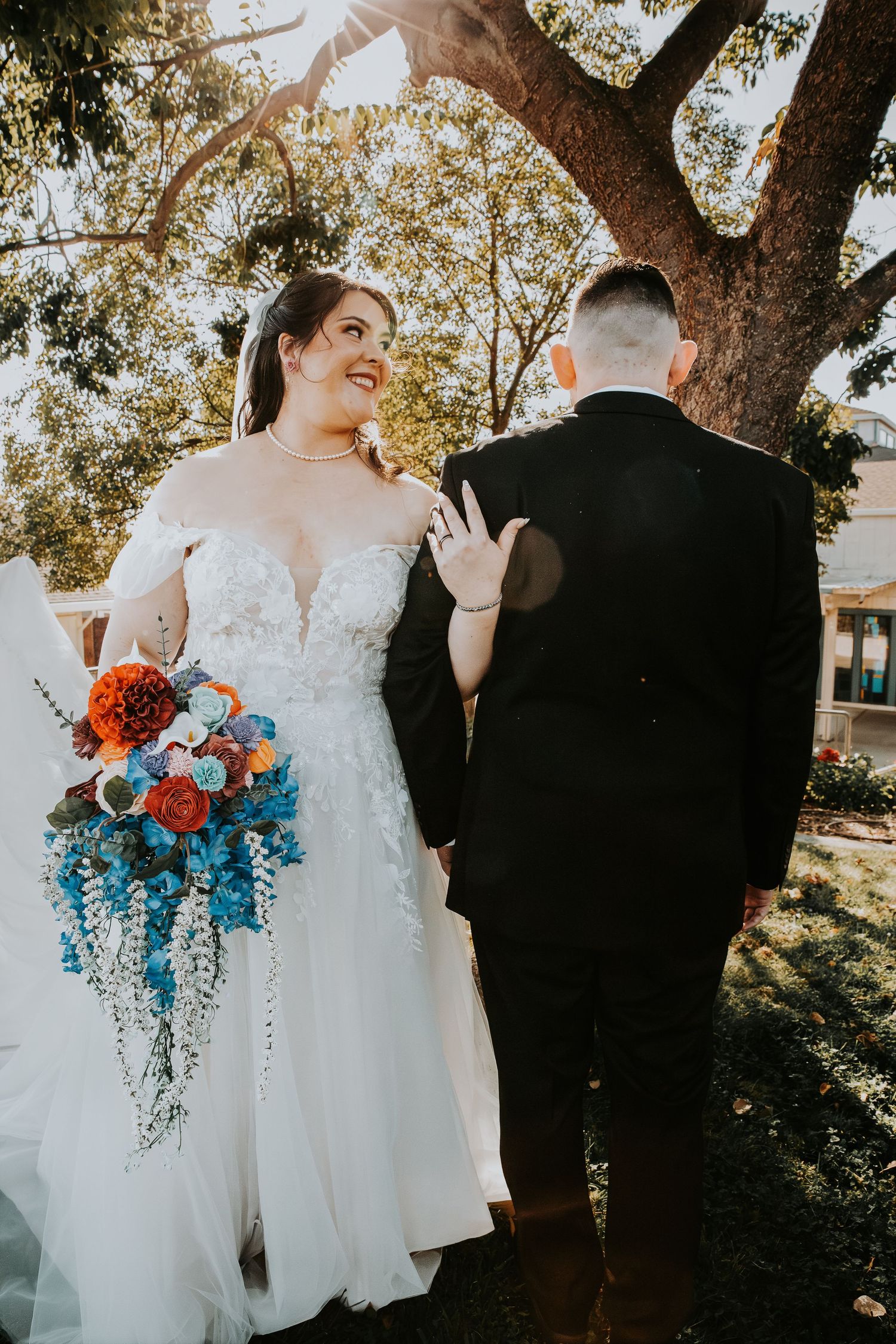 Bride in white lace off-shoulder gown holds colorful bouquet, smiling at groom in black suit outdoors.