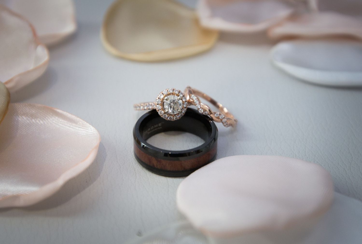 Rose gold halo diamond engagement ring with matching band and black wood inlay men's wedding ring on white petals.