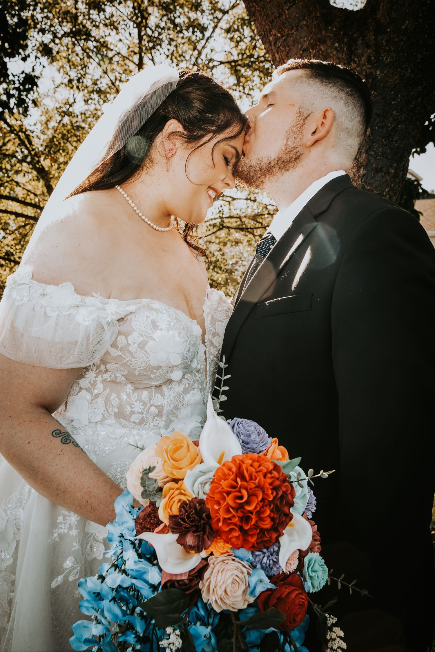 A bride and groom share a romantic kiss under a tree, with a vibrant bouquet of orange, blue, and pink flowers.