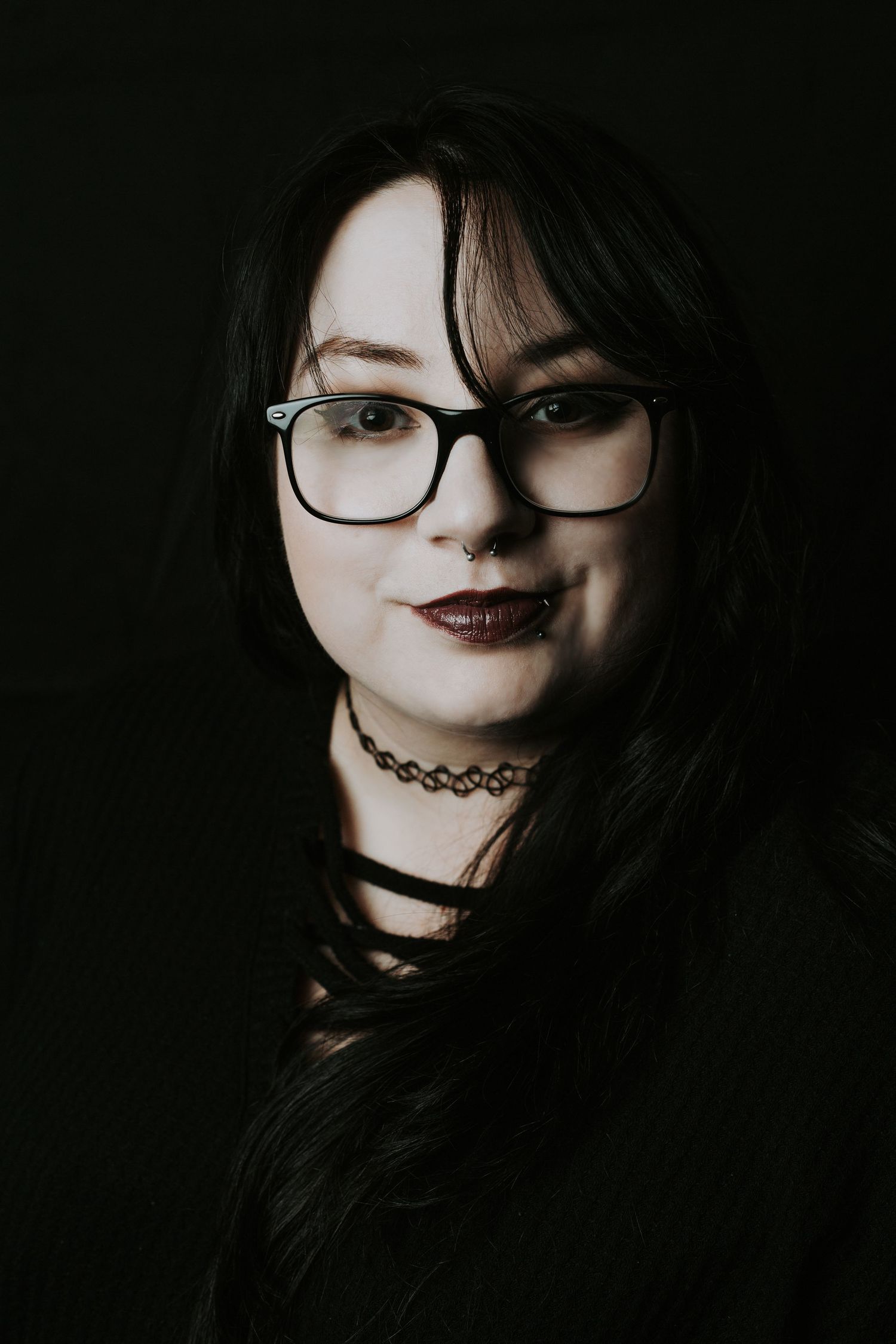 Young woman with black glasses, dark lipstick, nose ring, and choker necklace poses against a dark background.