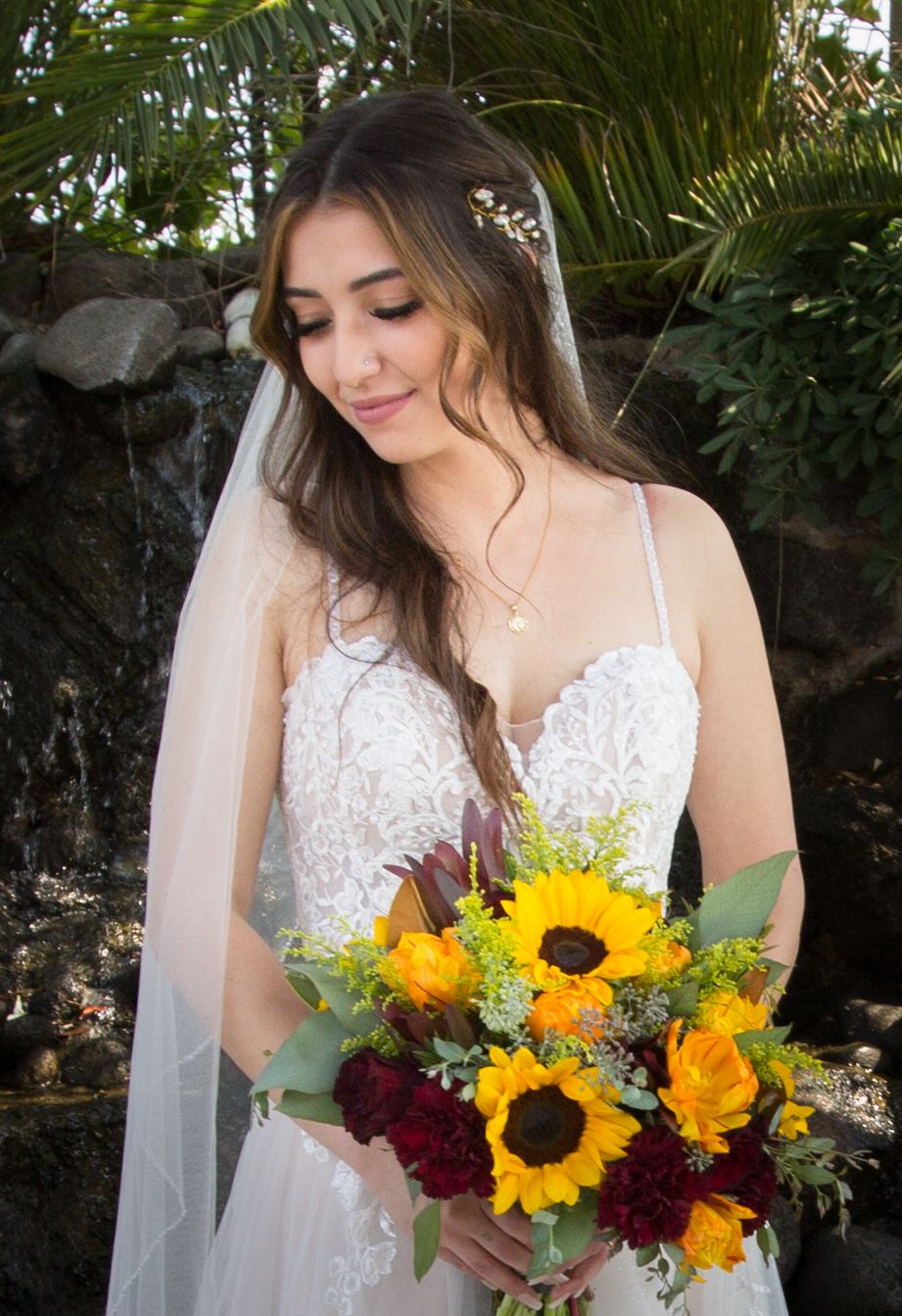 Bride in lace wedding dress and veil holds vibrant sunflower and red rose bouquet outdoors near a waterfall.