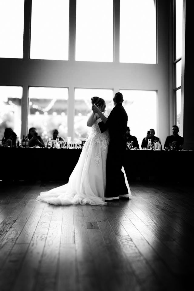 A black and white image of a bride and groom dancing on a wooden dance floor in a dimly lit venue.