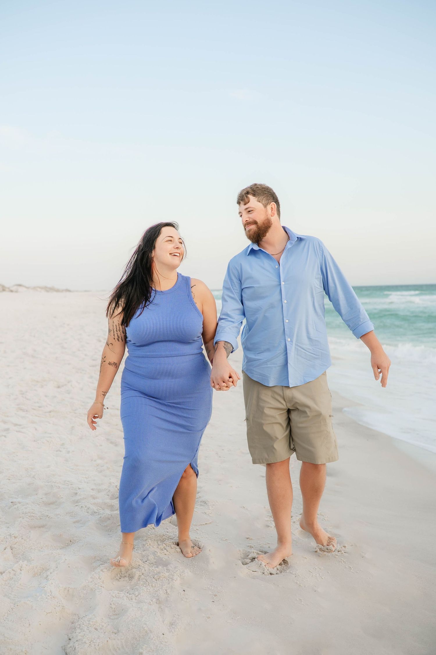 A couple walks hand in hand along the shoreline wearing coordinating blue outfits.