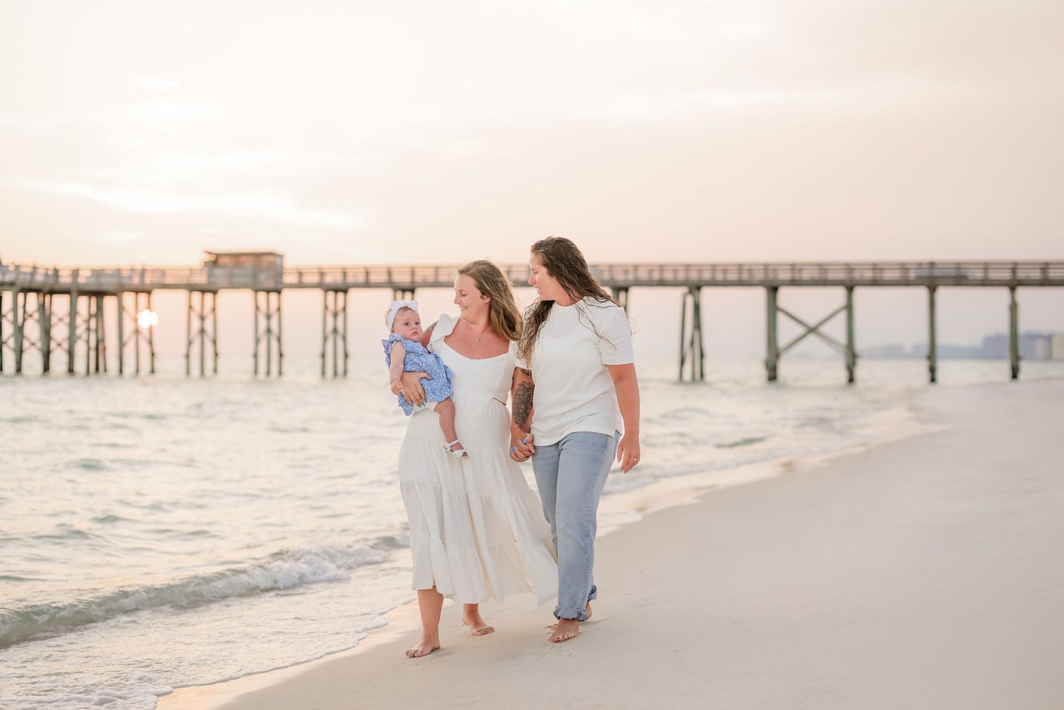 Three people stroll along a sandy beach during golden hour near a wooden pier.
