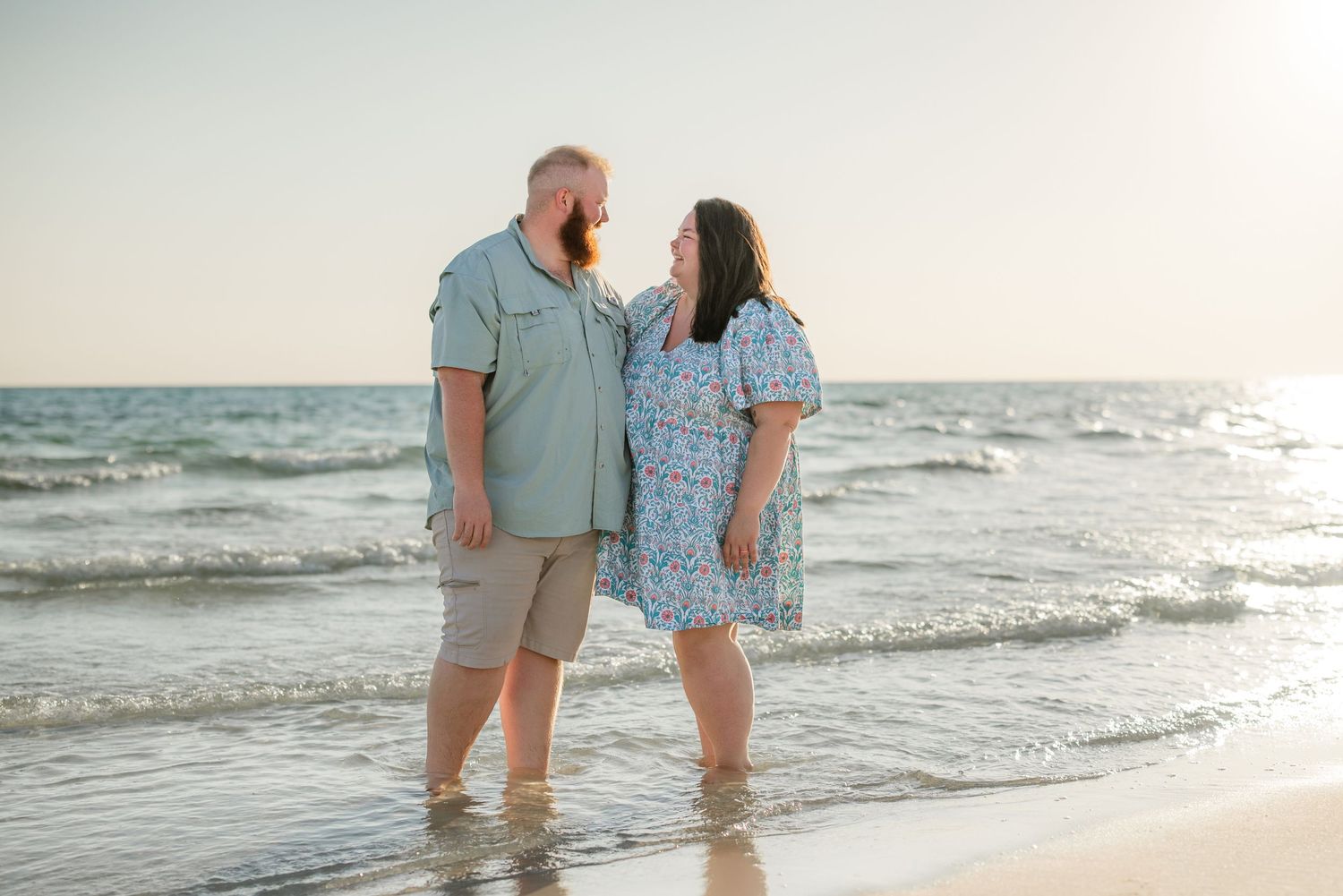 Couple standing together in gentle ocean waves at sunset wearing coordinated blue and khaki outfits.
