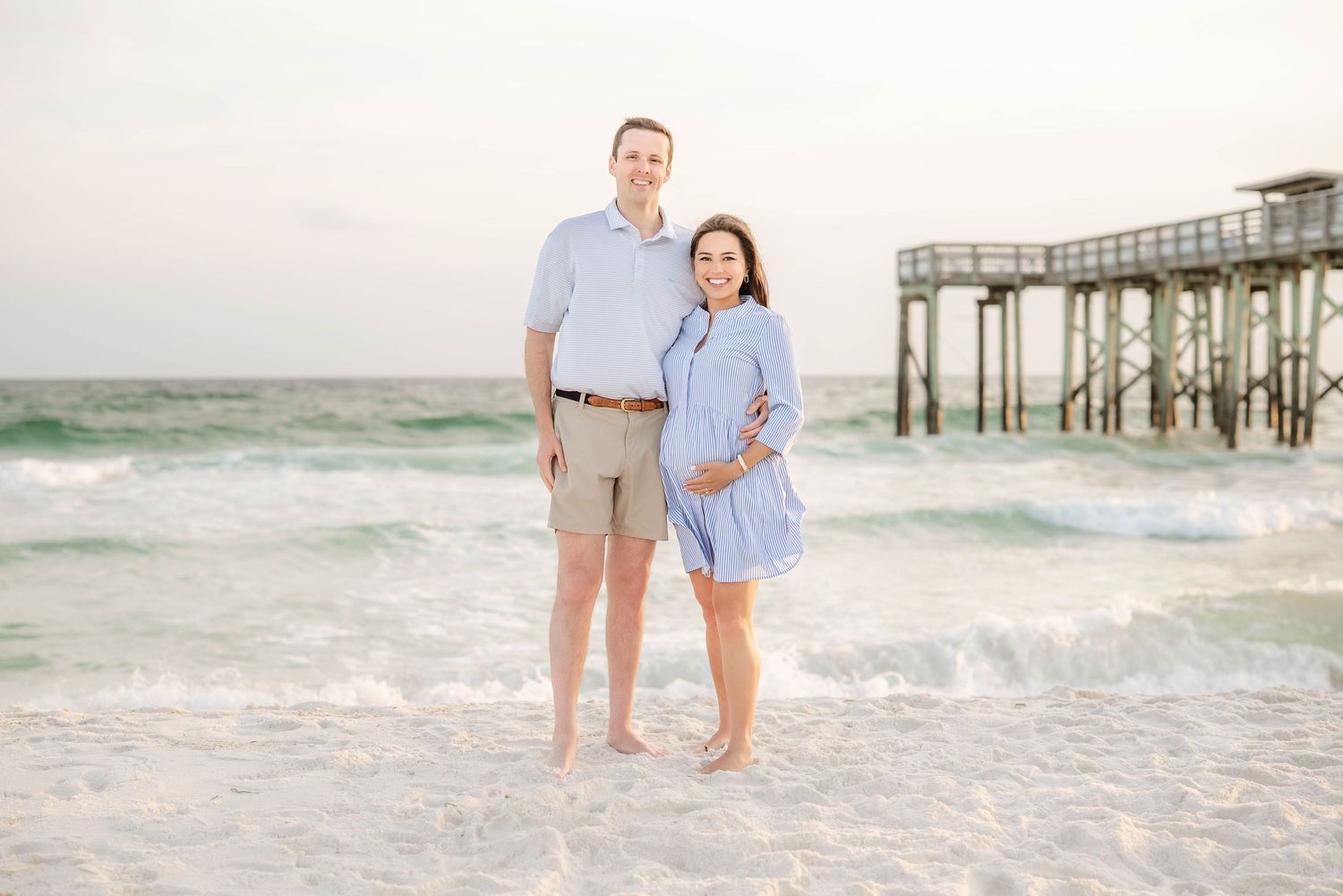 A couple shares a moment together on the beach with waves and a pier in the background.