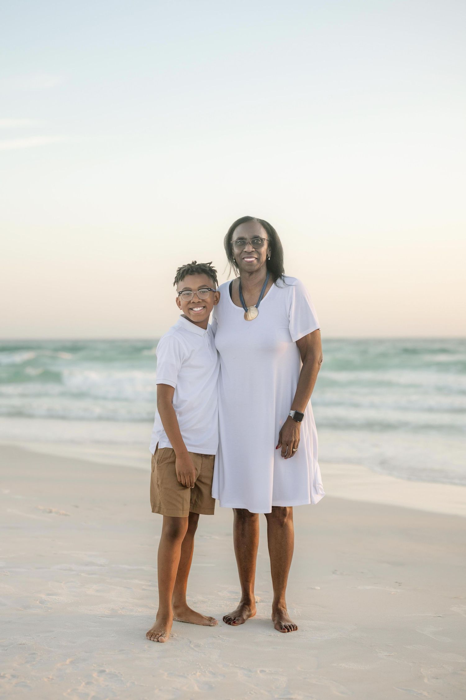 A woman and young boy in white outfits smile together on a sandy beach with ocean waves at sunset.