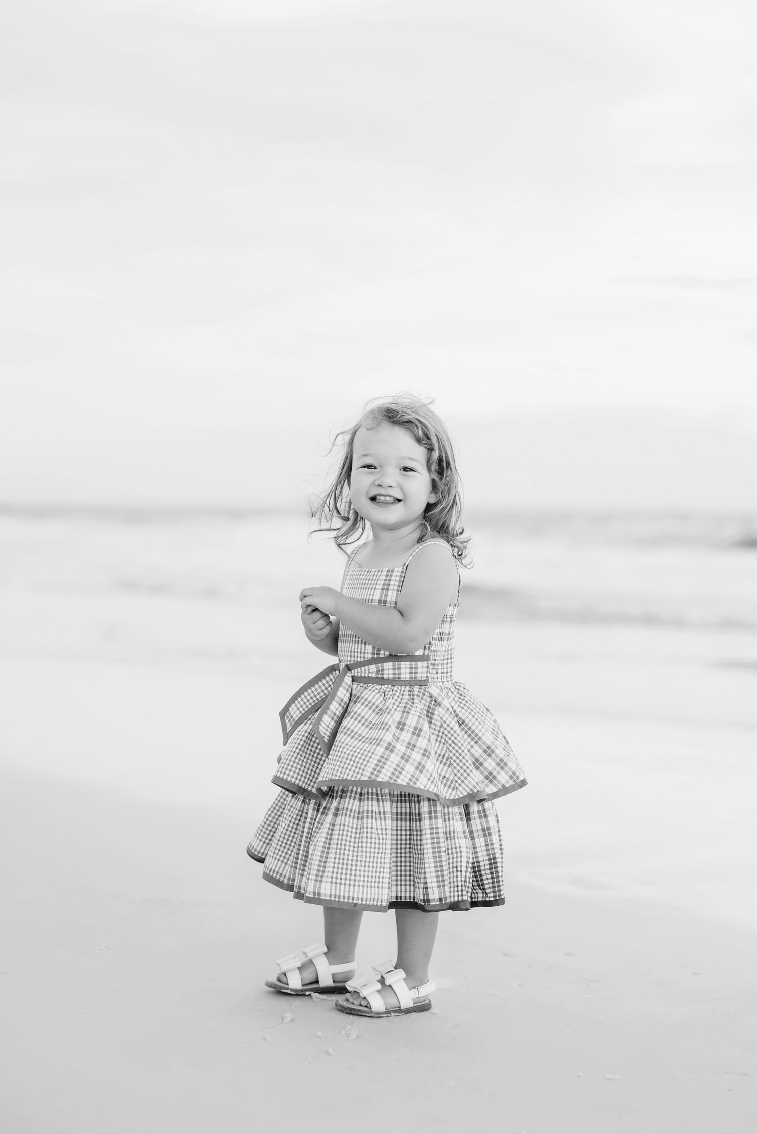 Black and white photo of child in gingham dress twirling on beach.