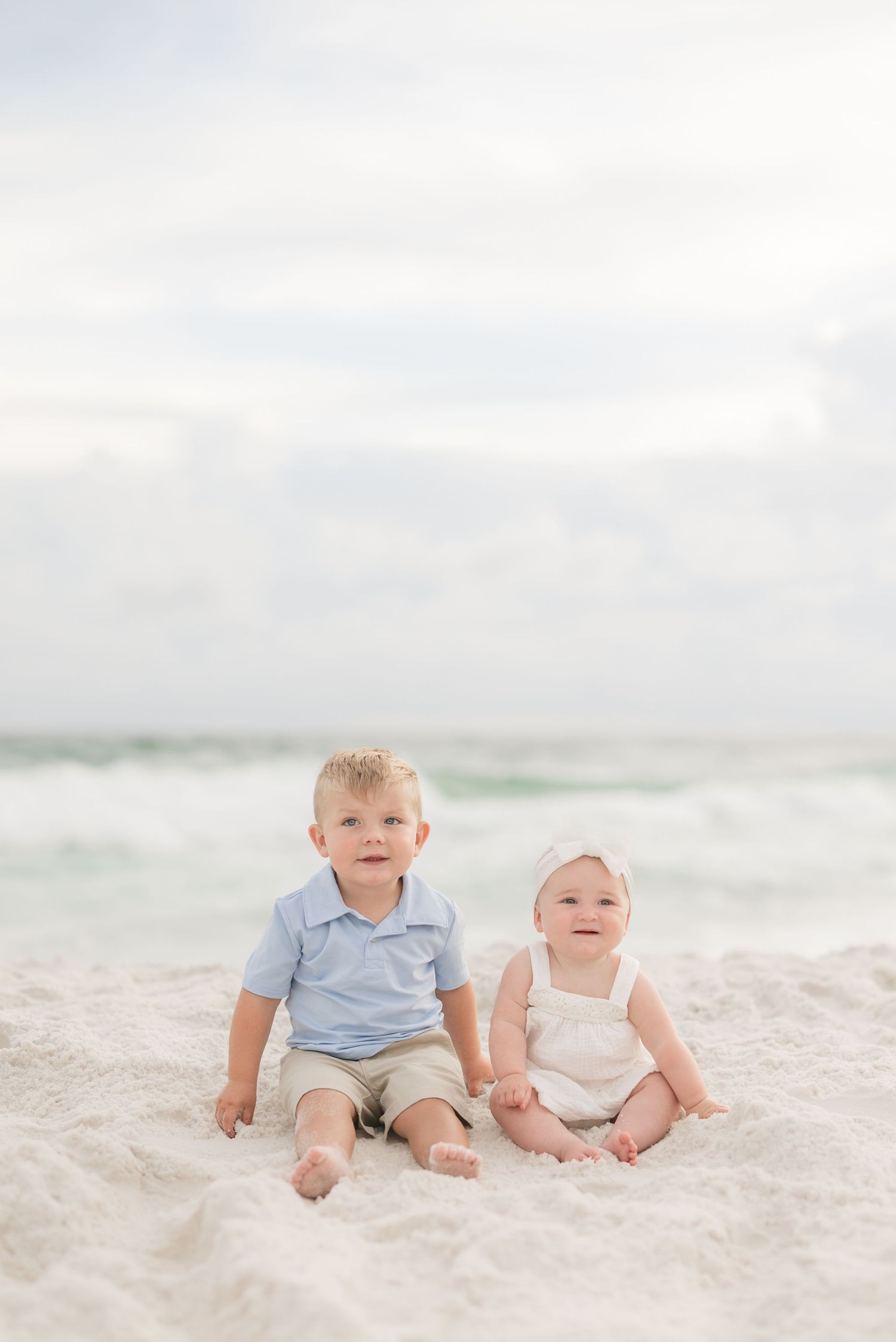 Two babies sitting together on a white sandy beach wearing light colored outfits.