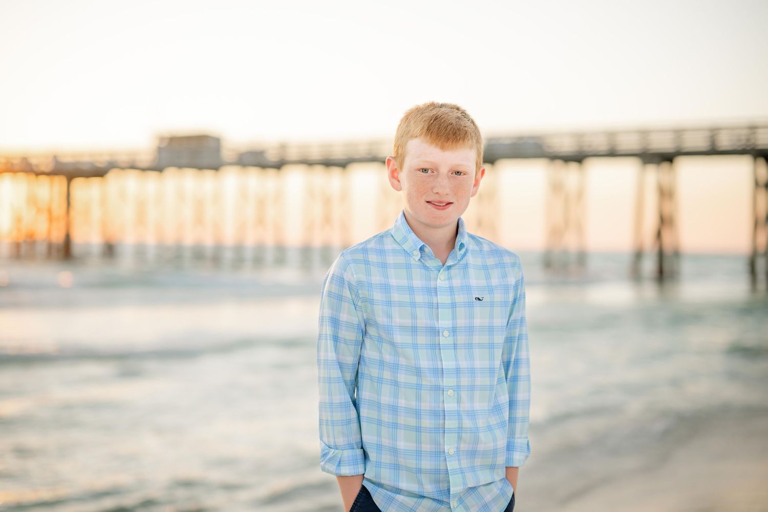 A person in a blue plaid shirt poses on a beach at sunset with a pier in the background.