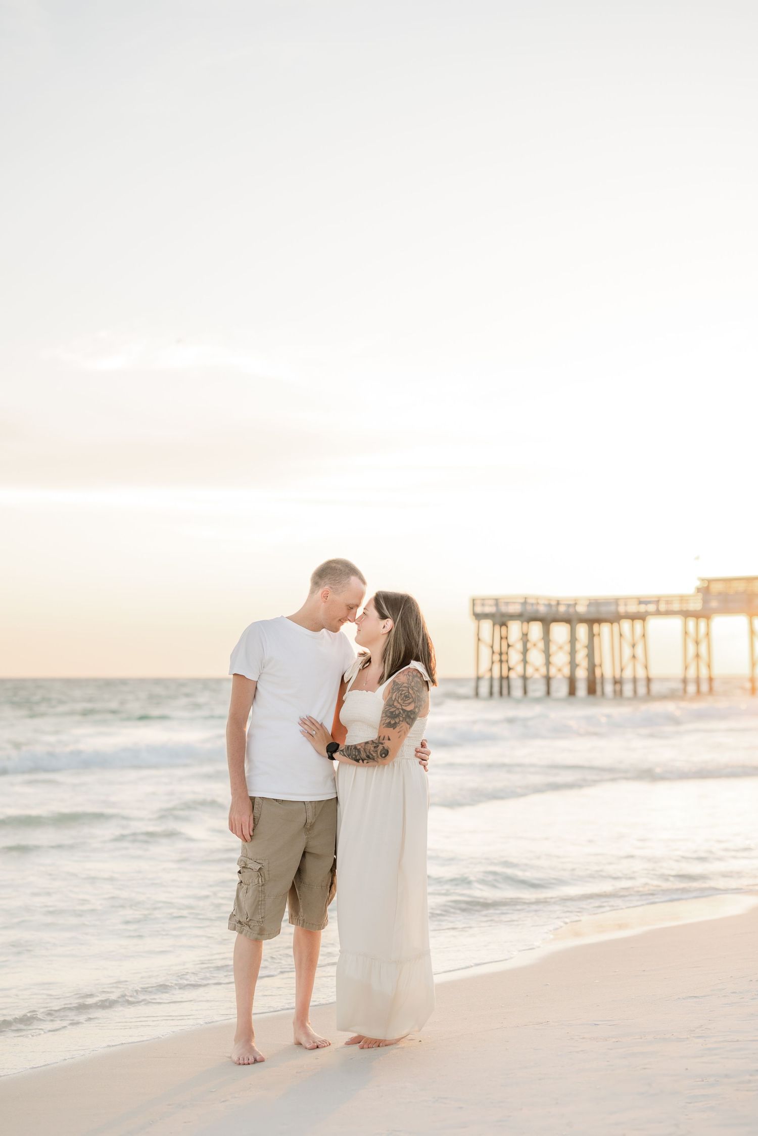 A couple walks hand in hand along a serene beach at sunset with a wooden pier in the background.